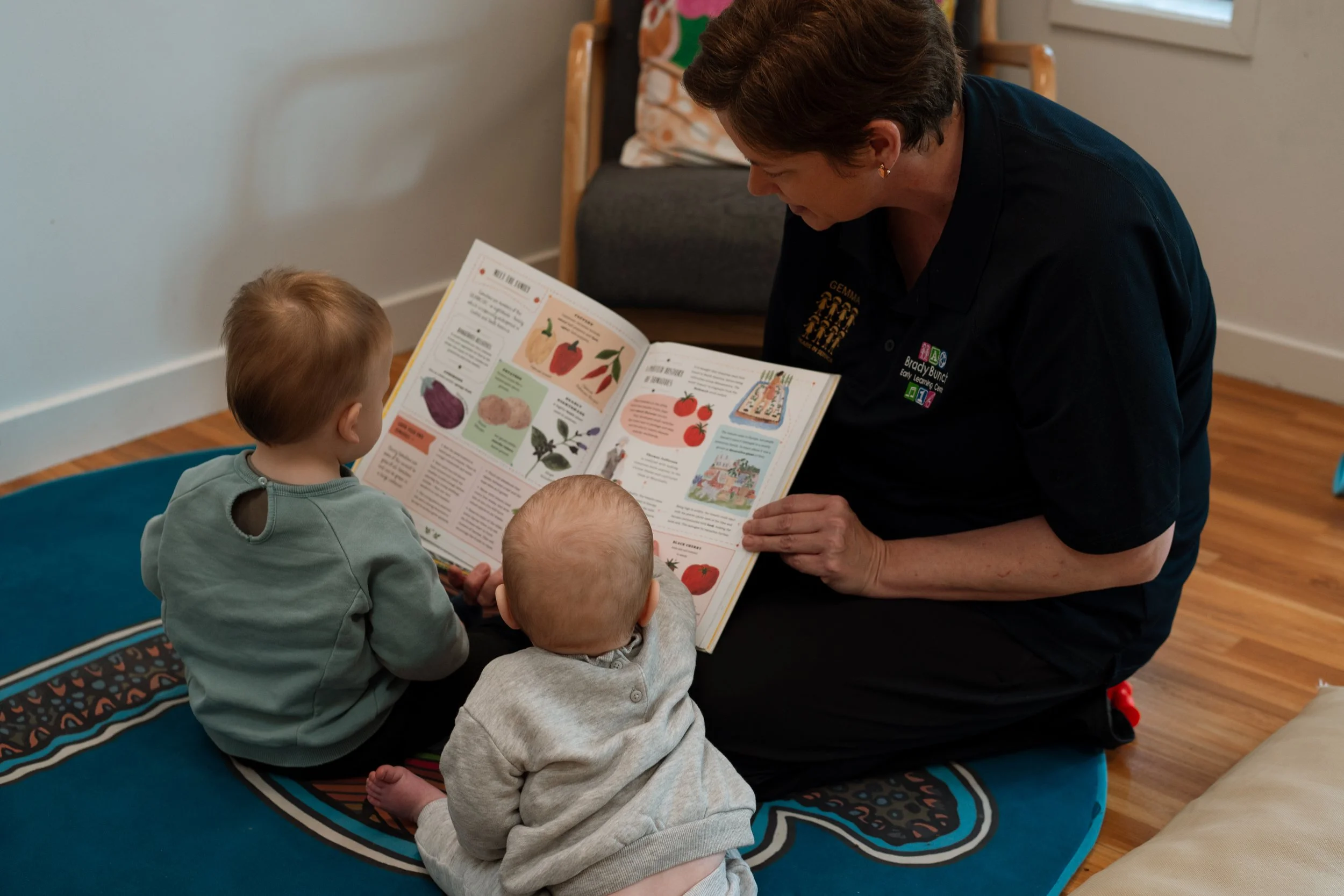 A woman reading a picture book to three young children seated on a blue rug in a room with wooden floors and white walls.