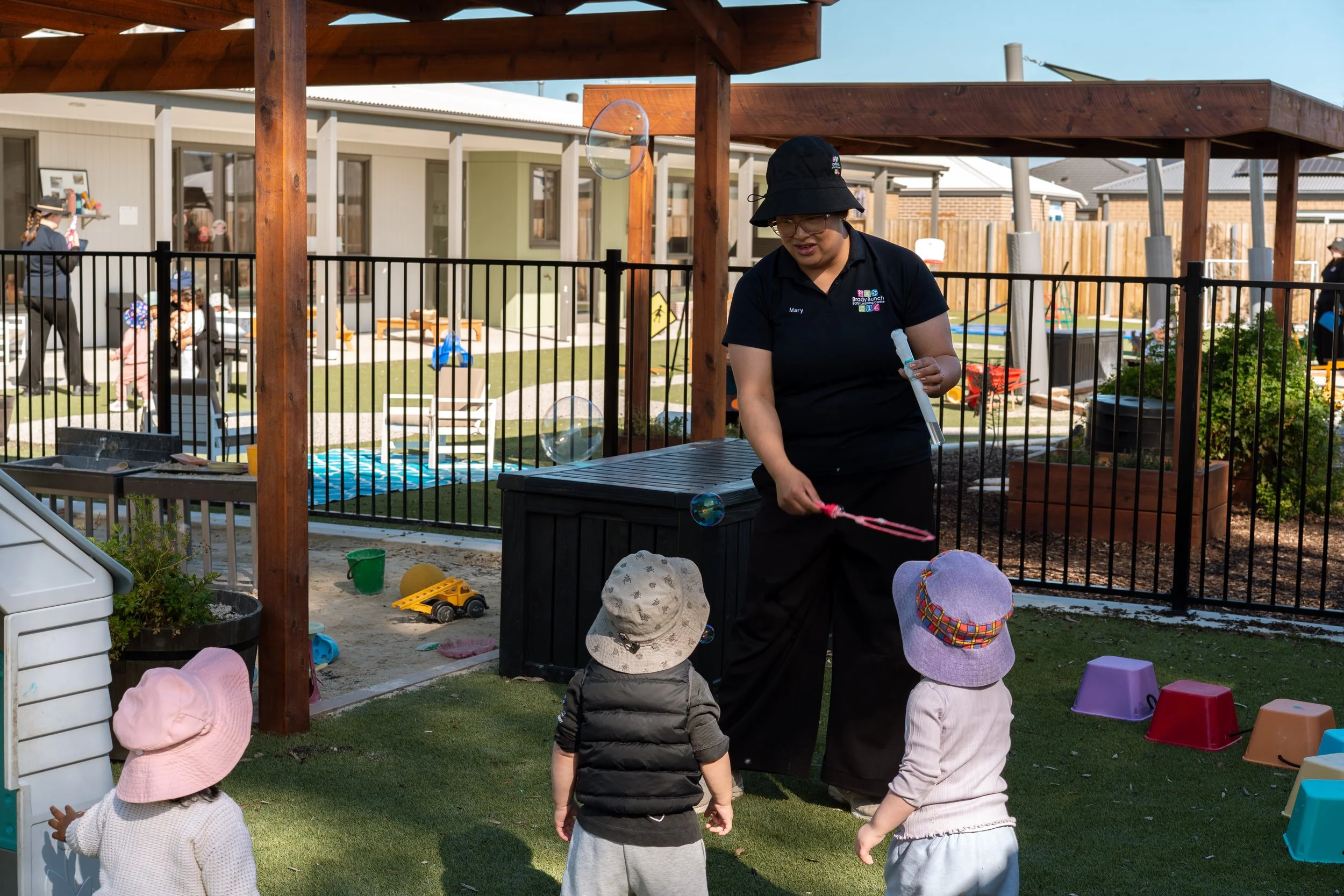 A woman is blowing bubbles while children watch outdoors in a playground area, with some toys and children wearing hats.