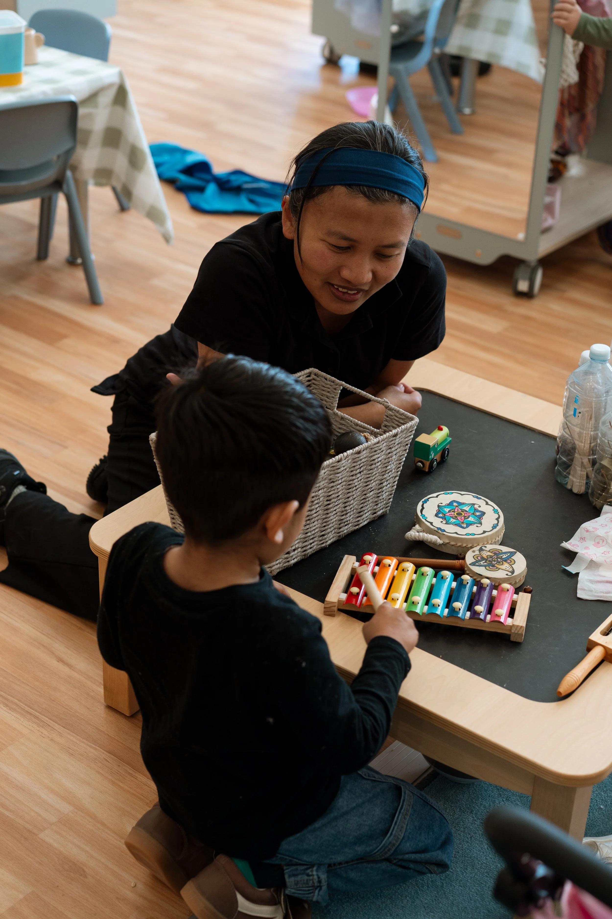 A woman and a young boy at a table playing with colorful toy xylophones and decorated wood slices. The woman is leaning over, smiling at the boy, who is holding a white mallet.