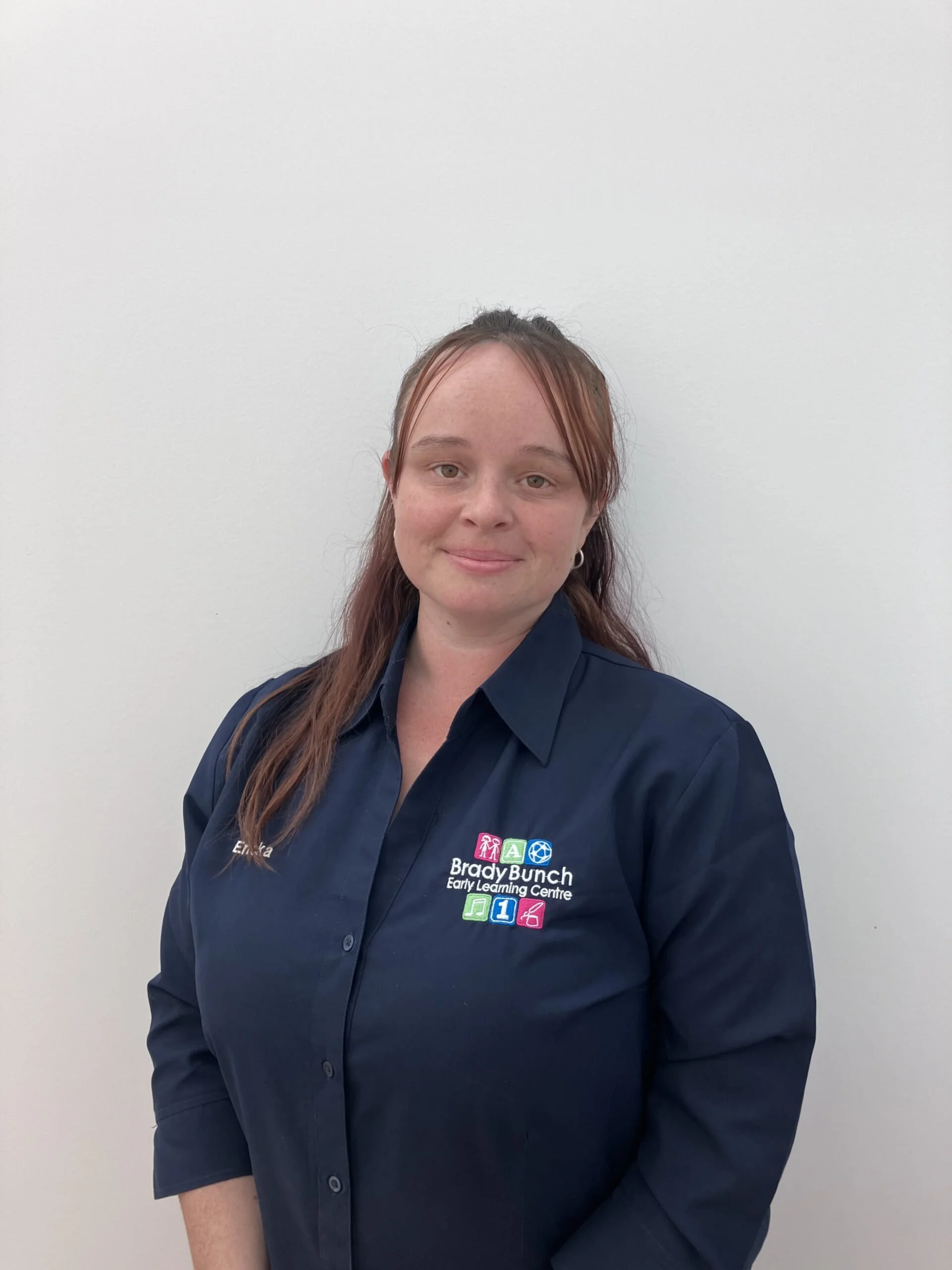 A woman with long red hair, wearing a dark blue collared shirt with a logo that reads 'Brady Bunch Early Learning Centre' and has colorful icons, standing against a plain white wall.
