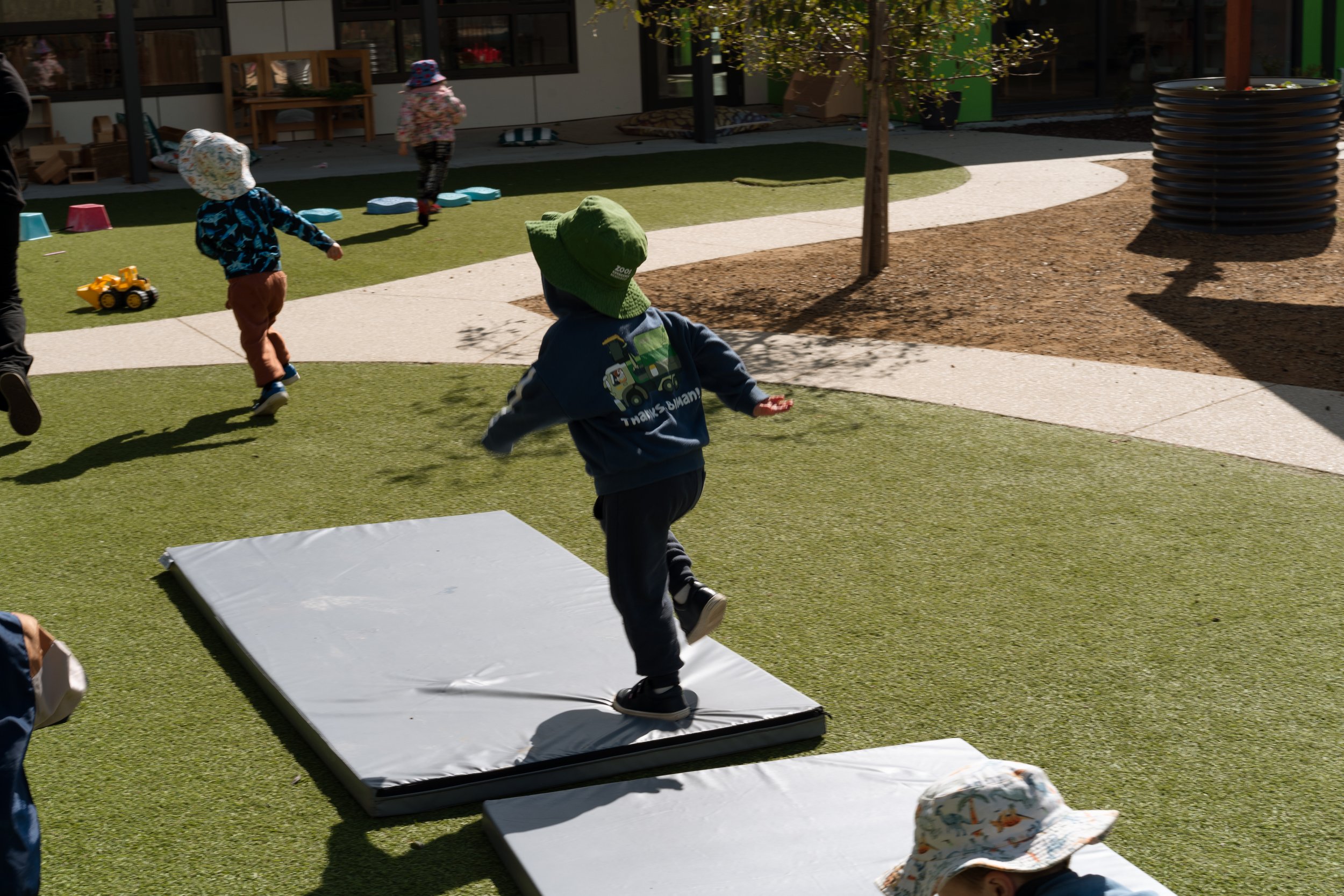 Children playing outdoors on a sunny day, with some children running and one stepping on a white mat on green artificial grass, surrounded by trees and a building in the background.