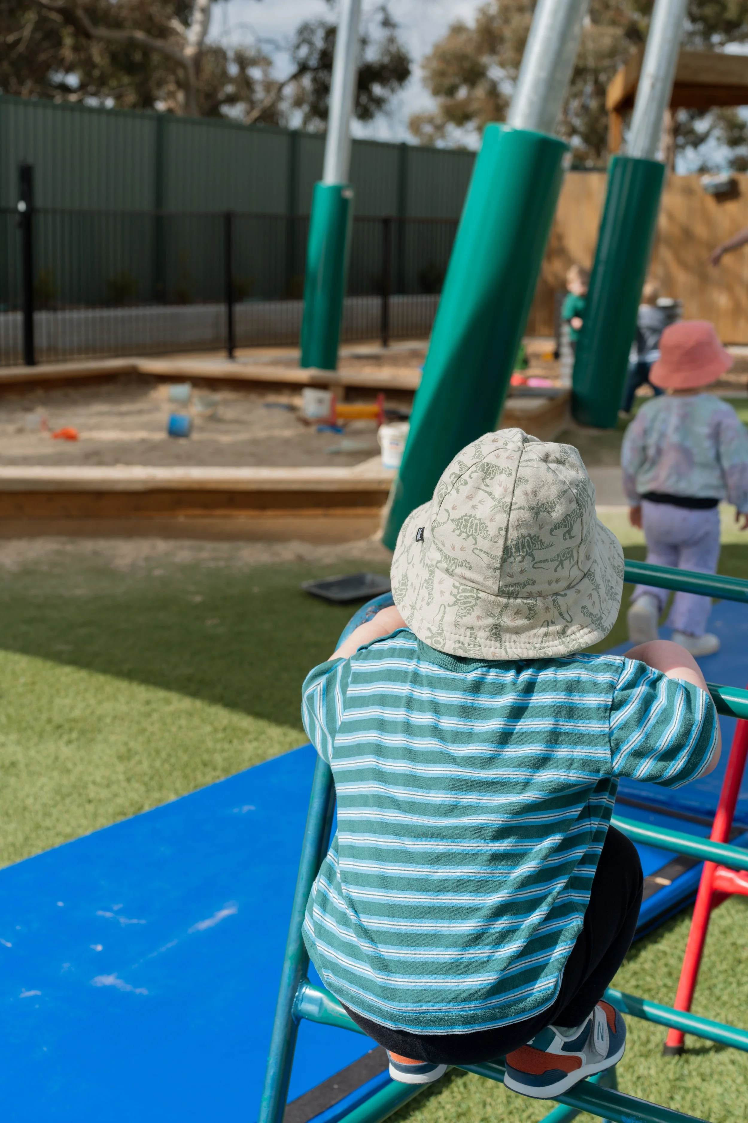 A young child in a striped shirt and black pants wearing sneakers, climbing on a playground structure near a sandbox, with other children playing in the background, in an outdoor playground area with a grassy ground, a fence, and trees.