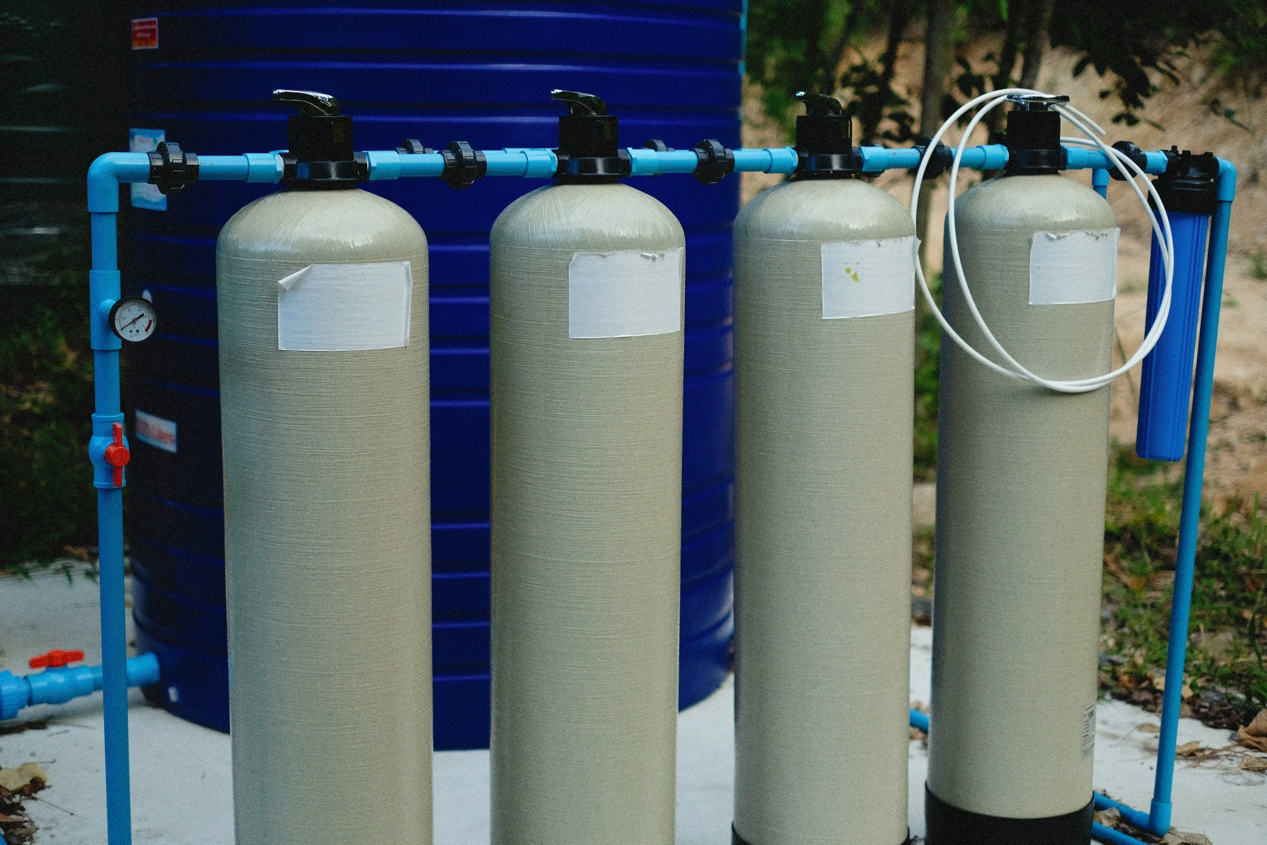 Water filtration system with four cylindrical tanks connected by blue pipes and black valve handles, with a pressure gauge and white tubing, set outdoors near a large blue tank and greenery.