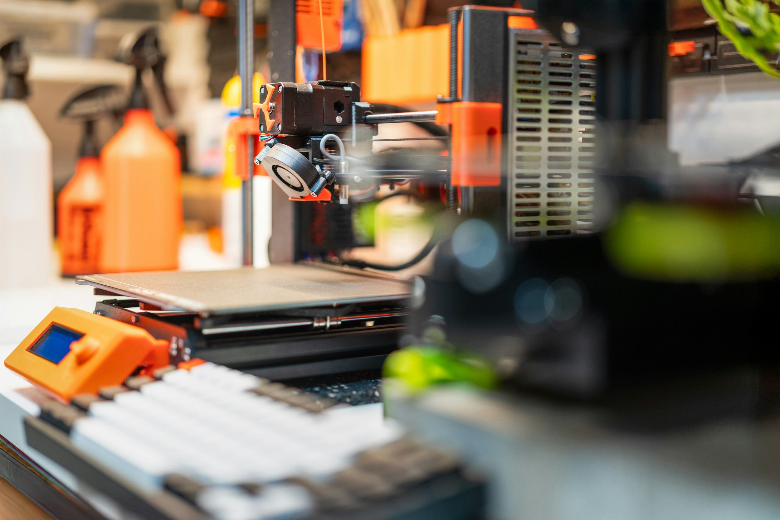 Close-up of a 3D printer with orange and black components, printing an object on a build plate in a workshop or lab setting, with various tools and supplies in the background.