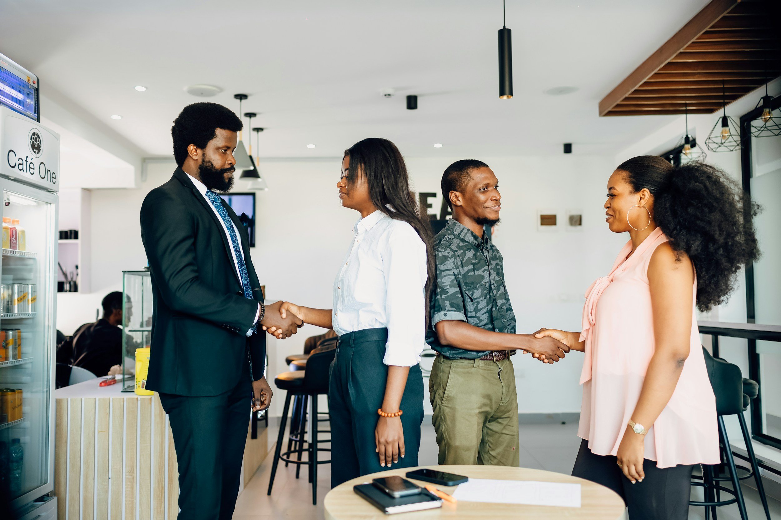 Two men and two women are shaking hands inside a modern coffee shop, engaging in a friendly greeting or meeting.