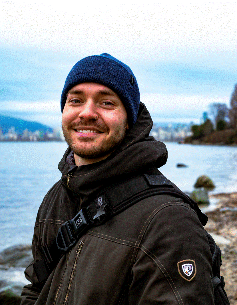 Young man smiling outdoors near water, wearing a blue beanie and dark jacket, with a cityscape and trees in the background.