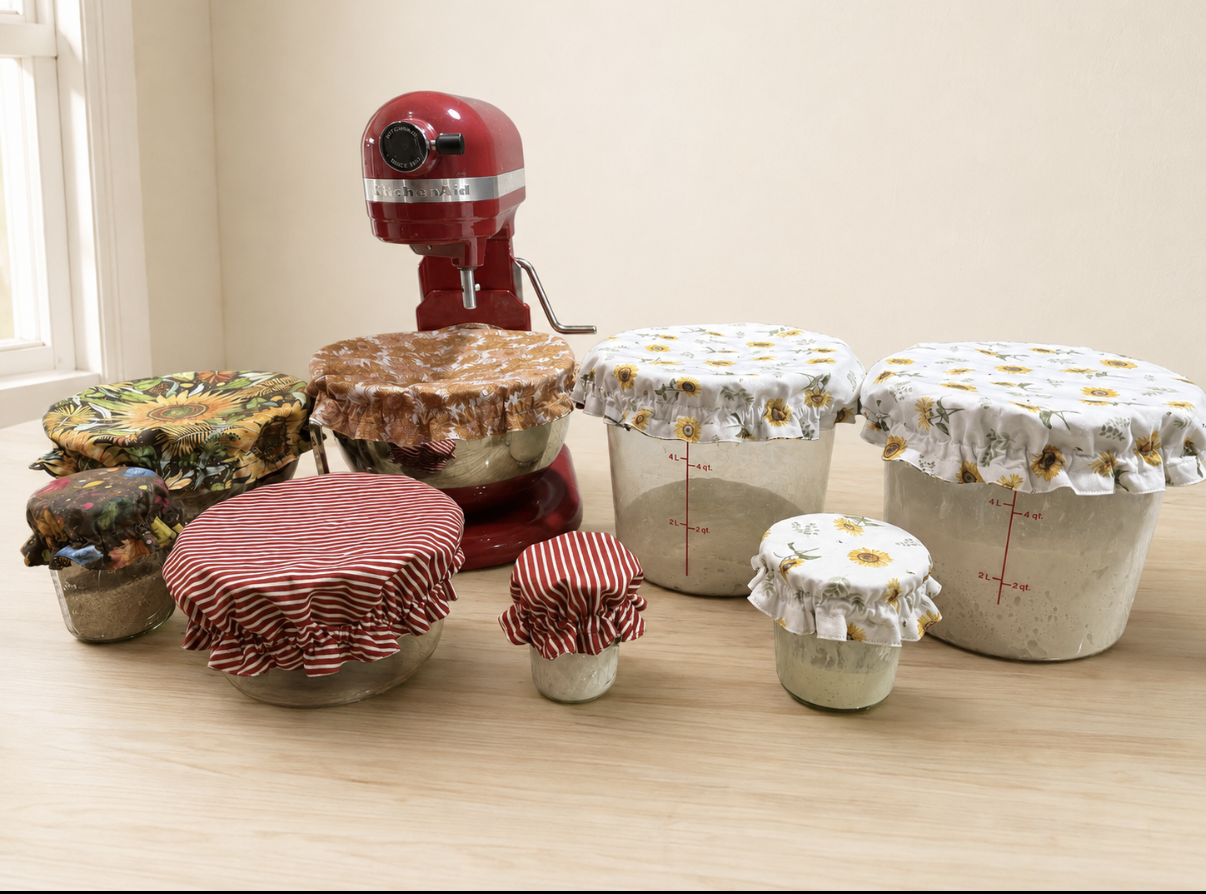 A red KitchenAid stand mixer with several glass jars and bowls covered with fabric kitchen towel covers.
