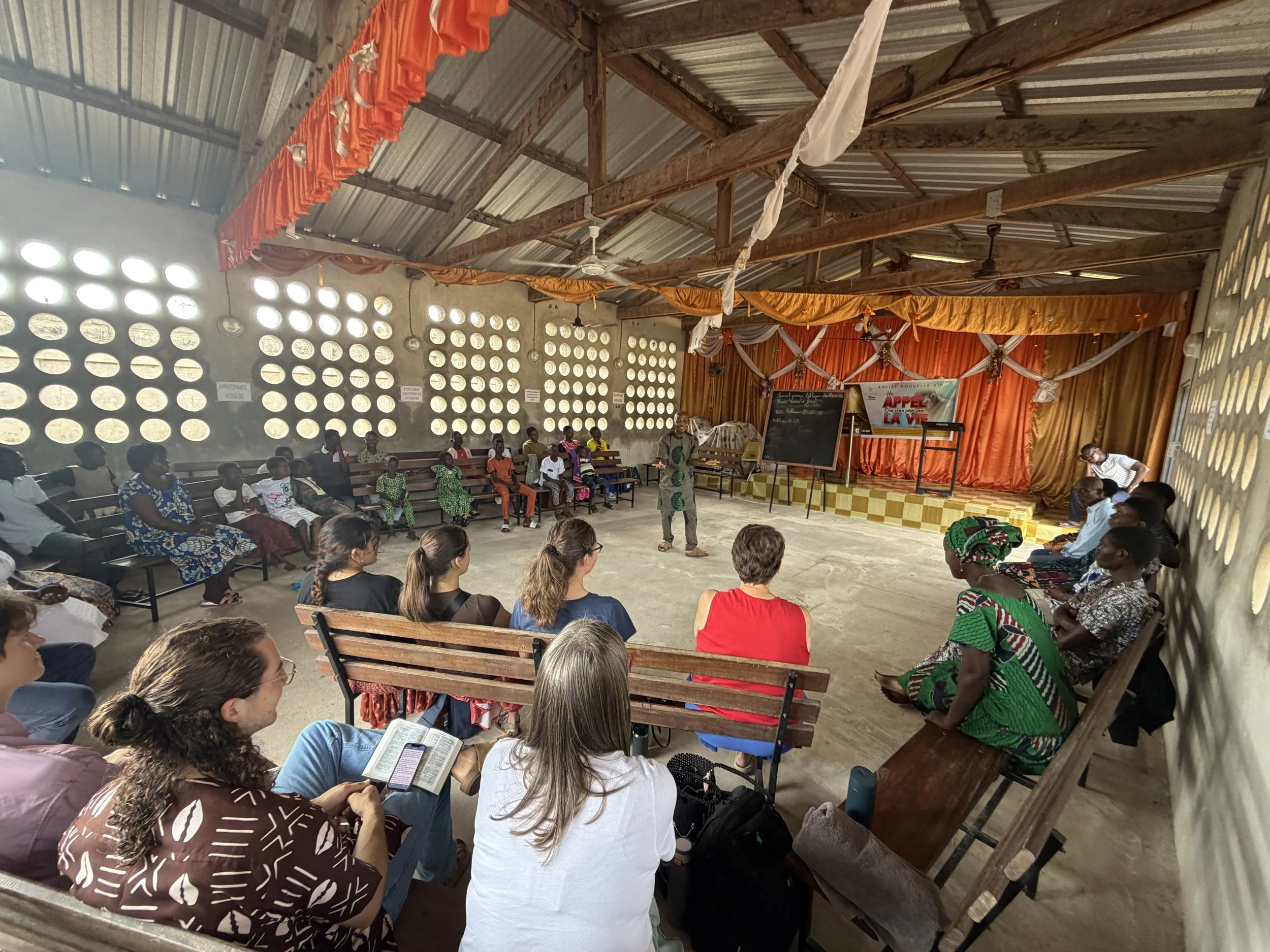 Inside a community center or meeting hall with a metal roof and open wall blocks, showing a group of people seated on benches and chairs, listening to a speaker standing near a blackboard and a stage decorated with orange and white drapes for an event or presentation.