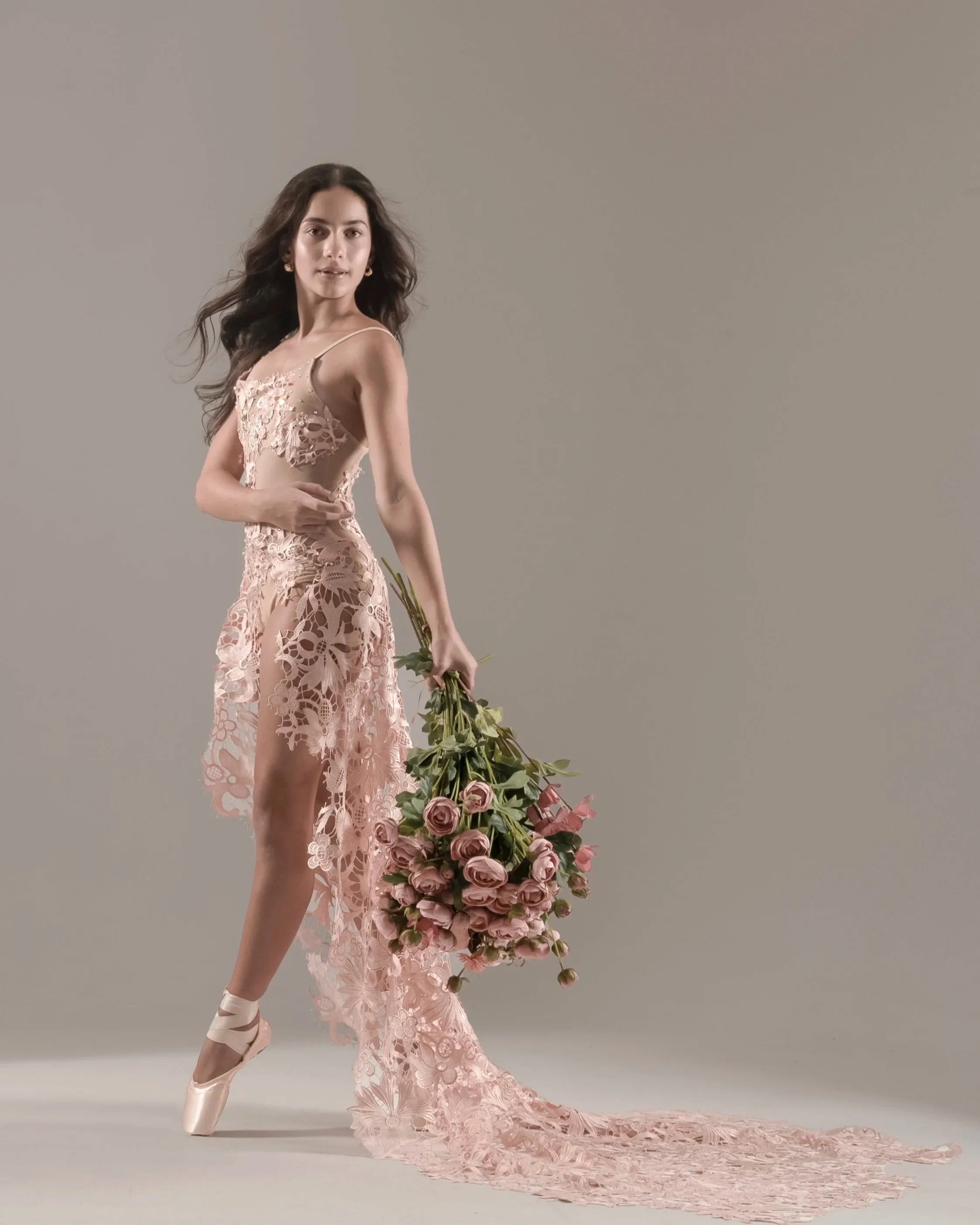 A young woman with long, wavy brown hair is wearing a nude-colored dress with intricate lace and floral embroidery. She is holding a large bouquet of roses and other flowers, and her left foot is pointed, showing off ballet shoes. She stands in front of a plain gray background.