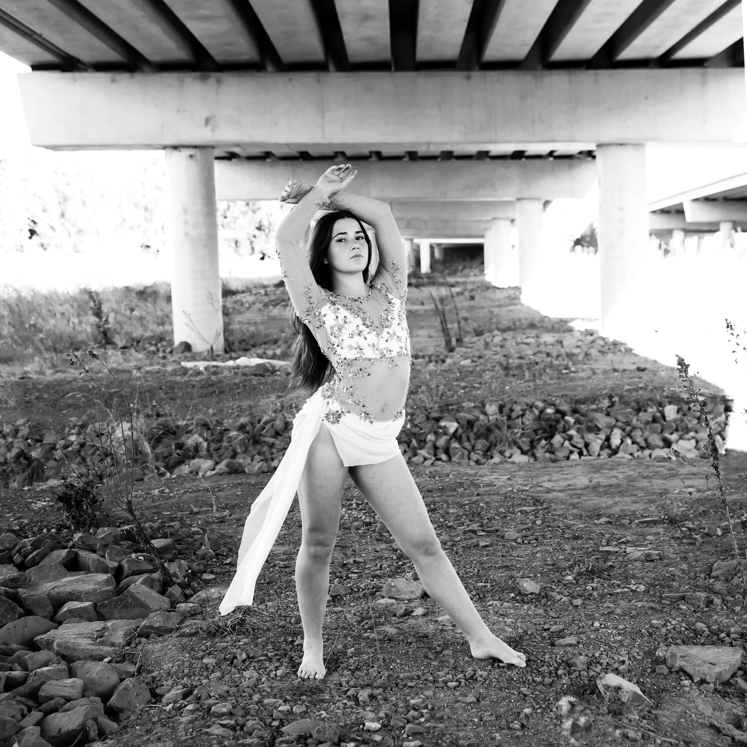 A woman with long hair posing under a bridge in black and white, wearing a sheer, embellished top and a high-slit skirt, with her arms raised above her head.