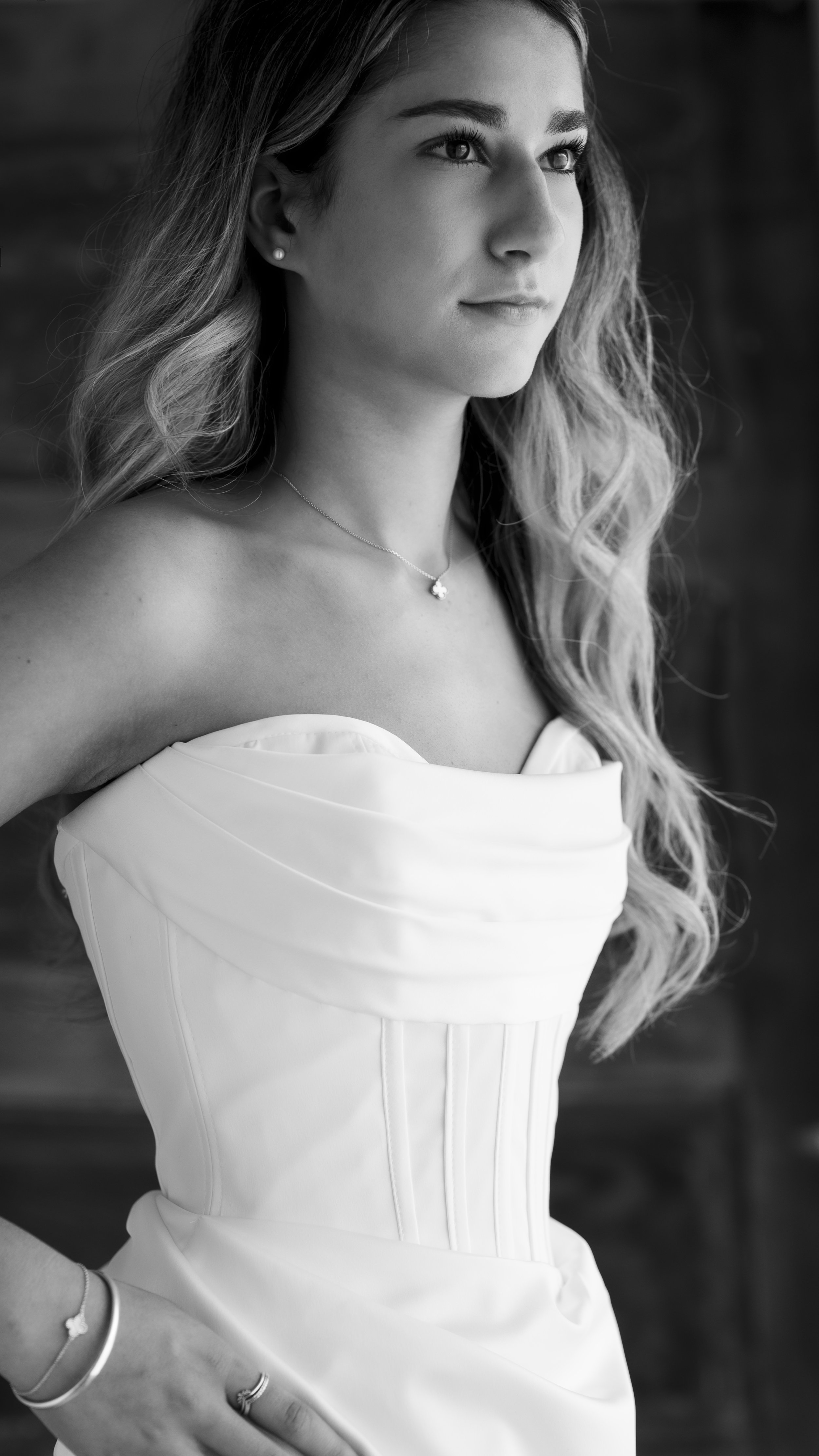 Black and white portrait of a young woman with long, wavy hair wearing a strapless dress, necklace, earrings, and bracelets.
