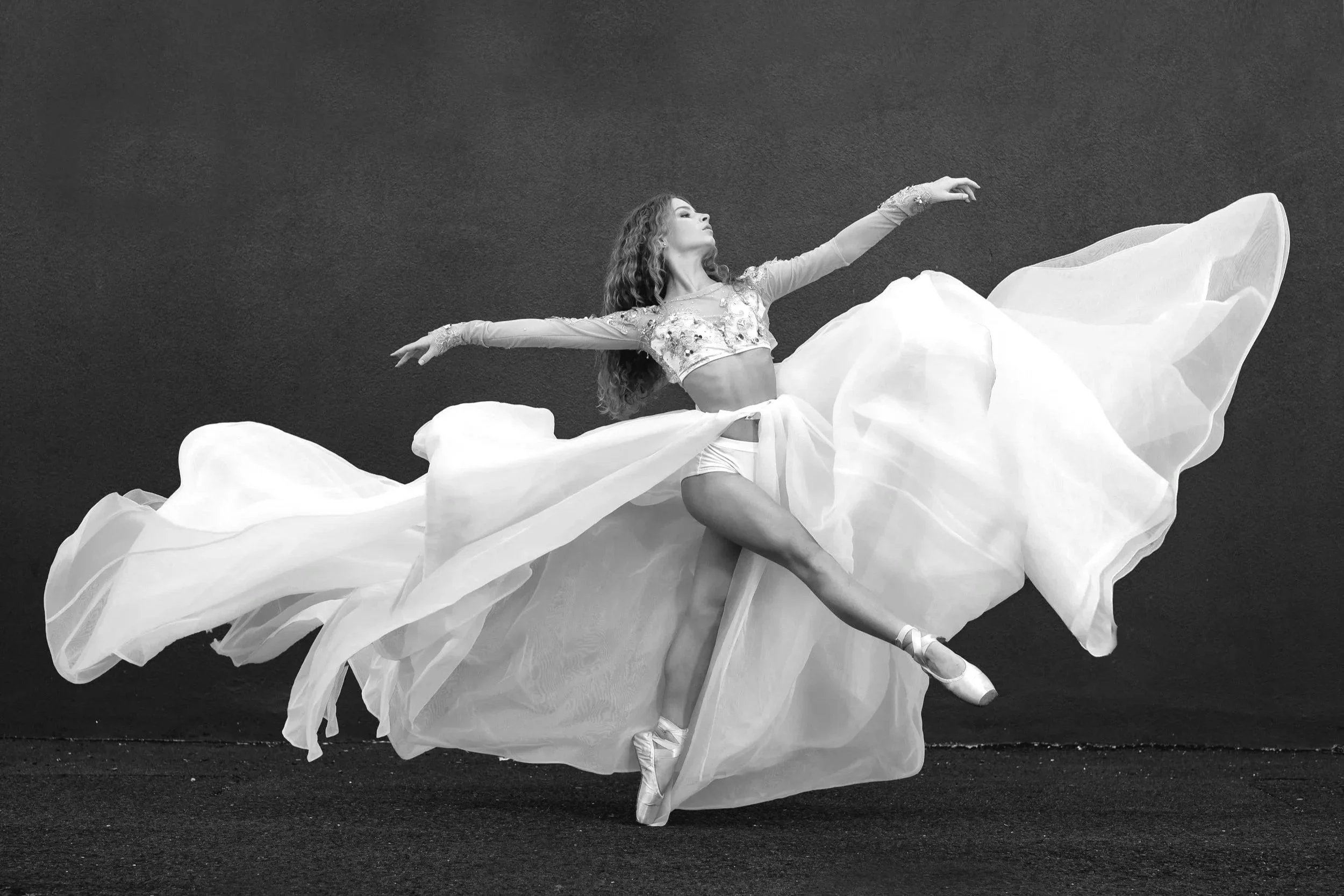 A female ballet dancer in a flowing dress performing a dance pose against a dark background.