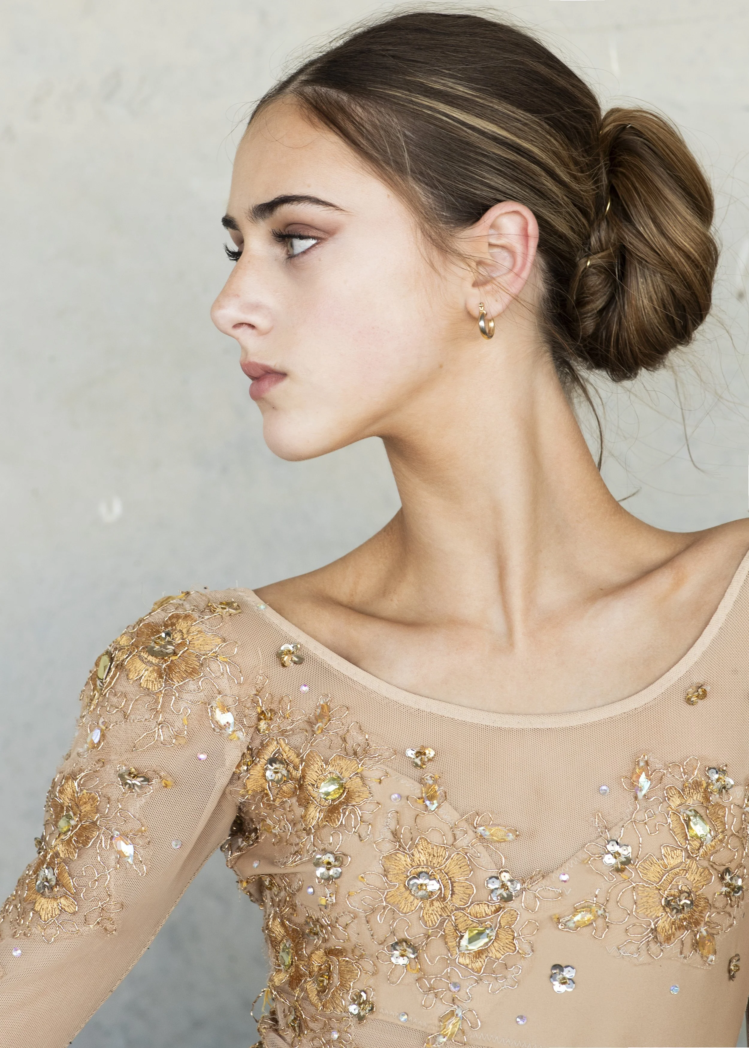 A woman with light skin and brown hair styled in a low bun, wearing a beige, sheer dress with gold floral embroidery and rhinestones, earrings, looking to her left against a neutral background.