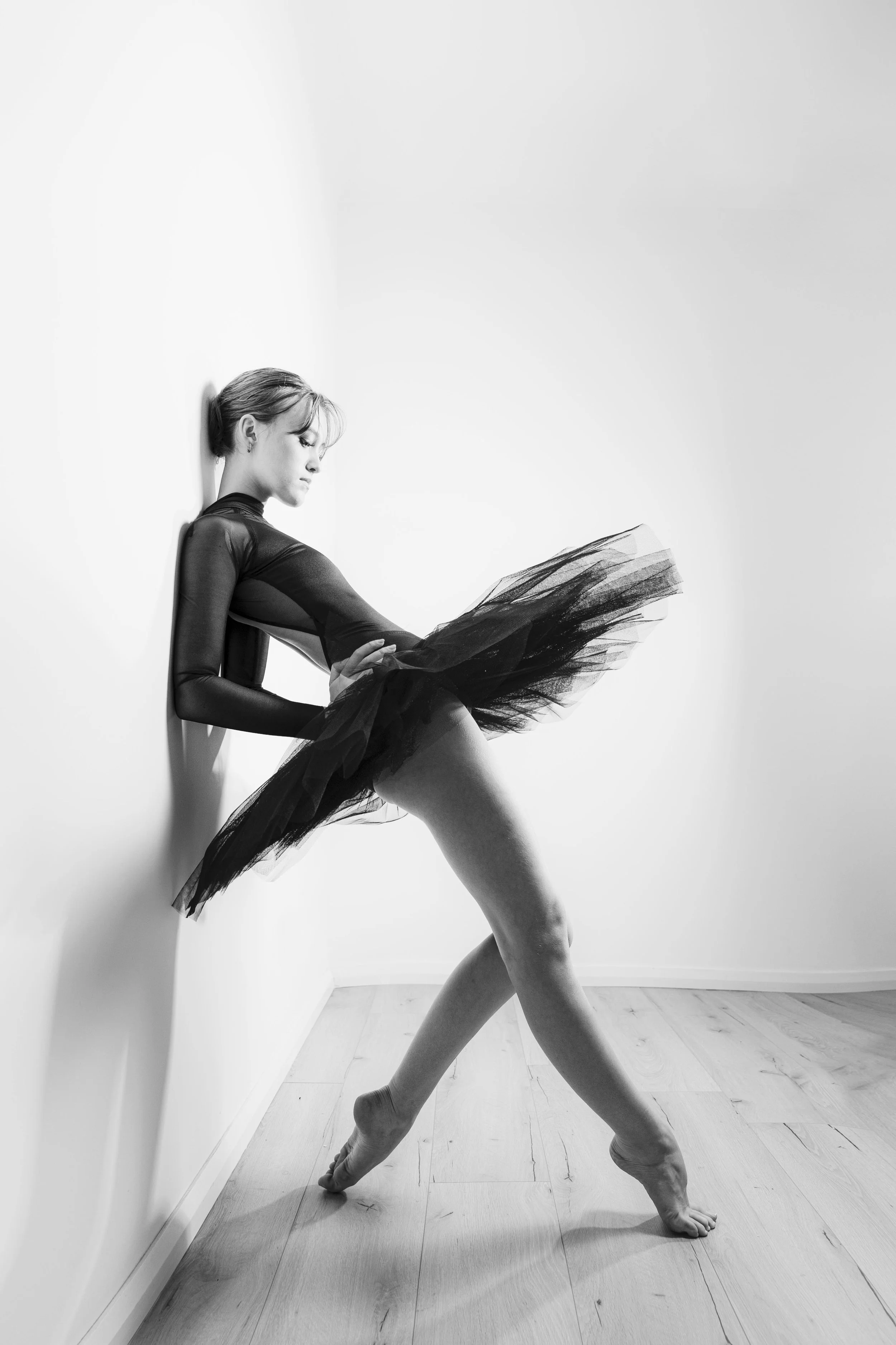 A ballerina leaning against a white wall in a dance studio, wearing a black tulle tutu and a long-sleeved black leotard, with one leg extended forward and the other leg bent, supporting her weight with her foot flat on the floor.