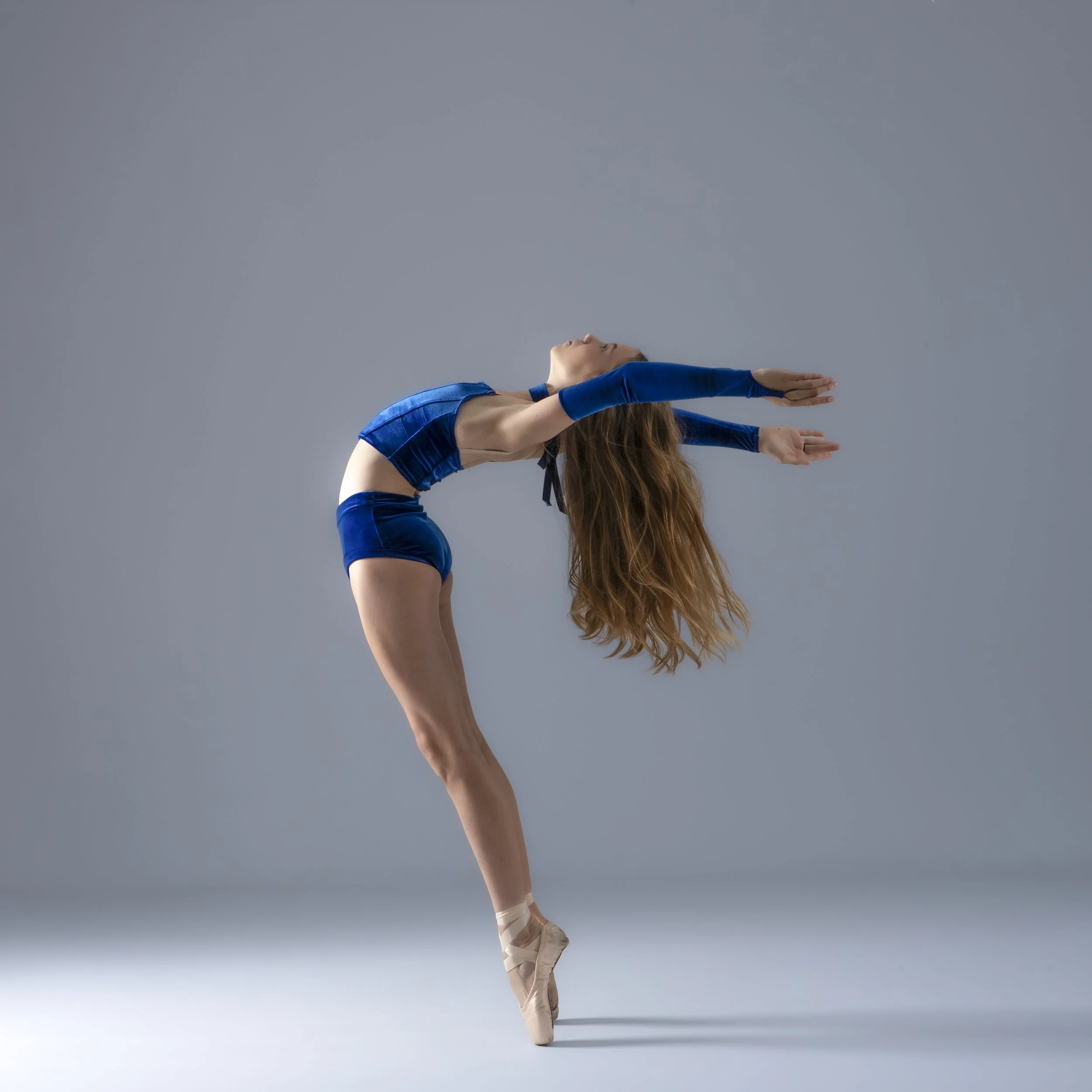 A dancer in a blue costume performing a pose on pointe shoes against a neutral background.