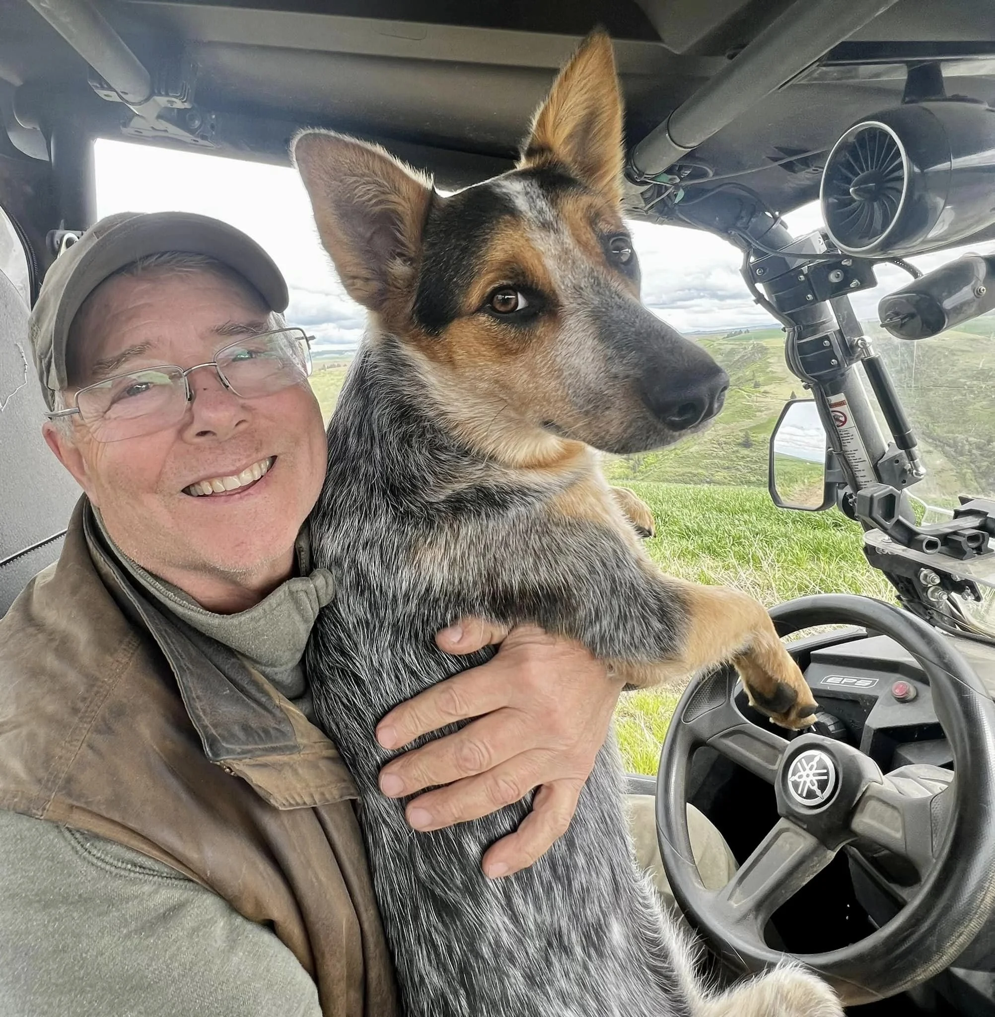 A man holding a dog in the cockpit of an aircraft, with the landscape visible outside through the windshield.