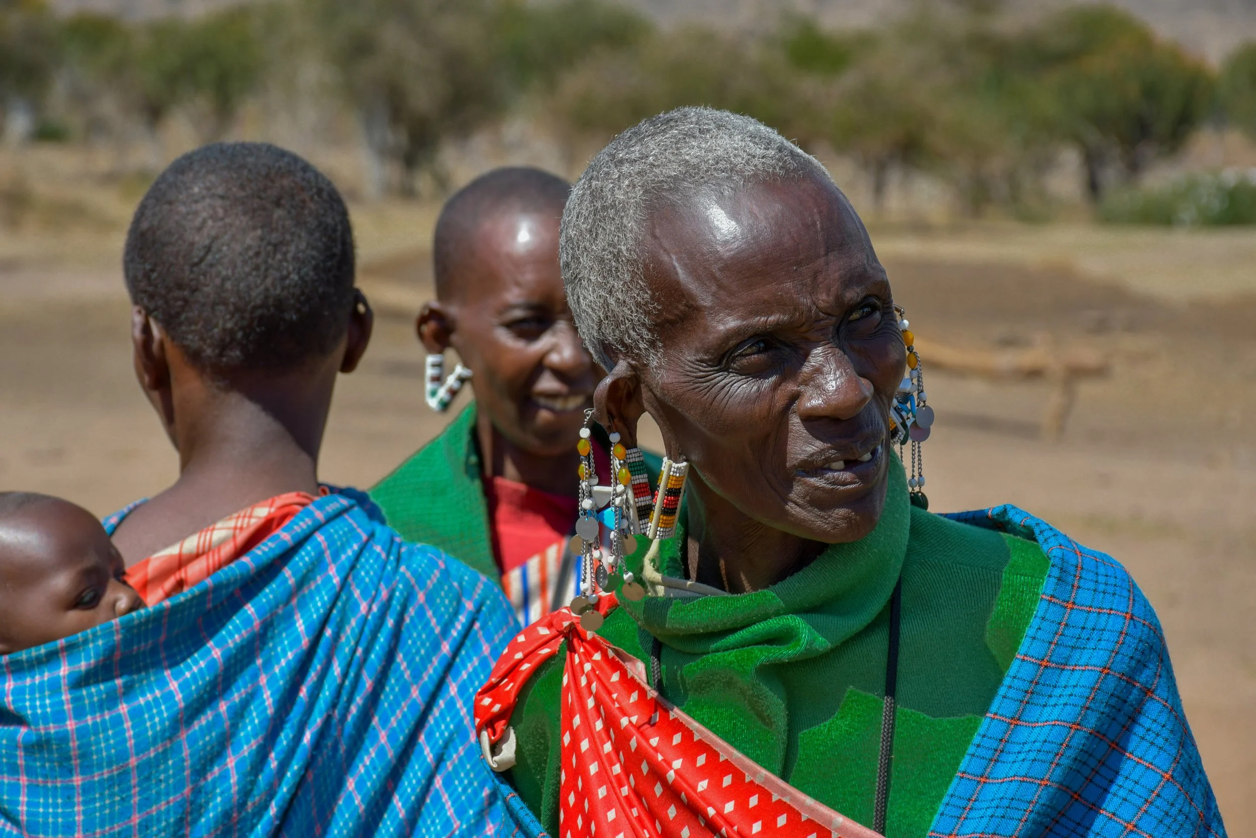 A woman wearing colorful beaded earrings and patterned clothing, with community members nearby.