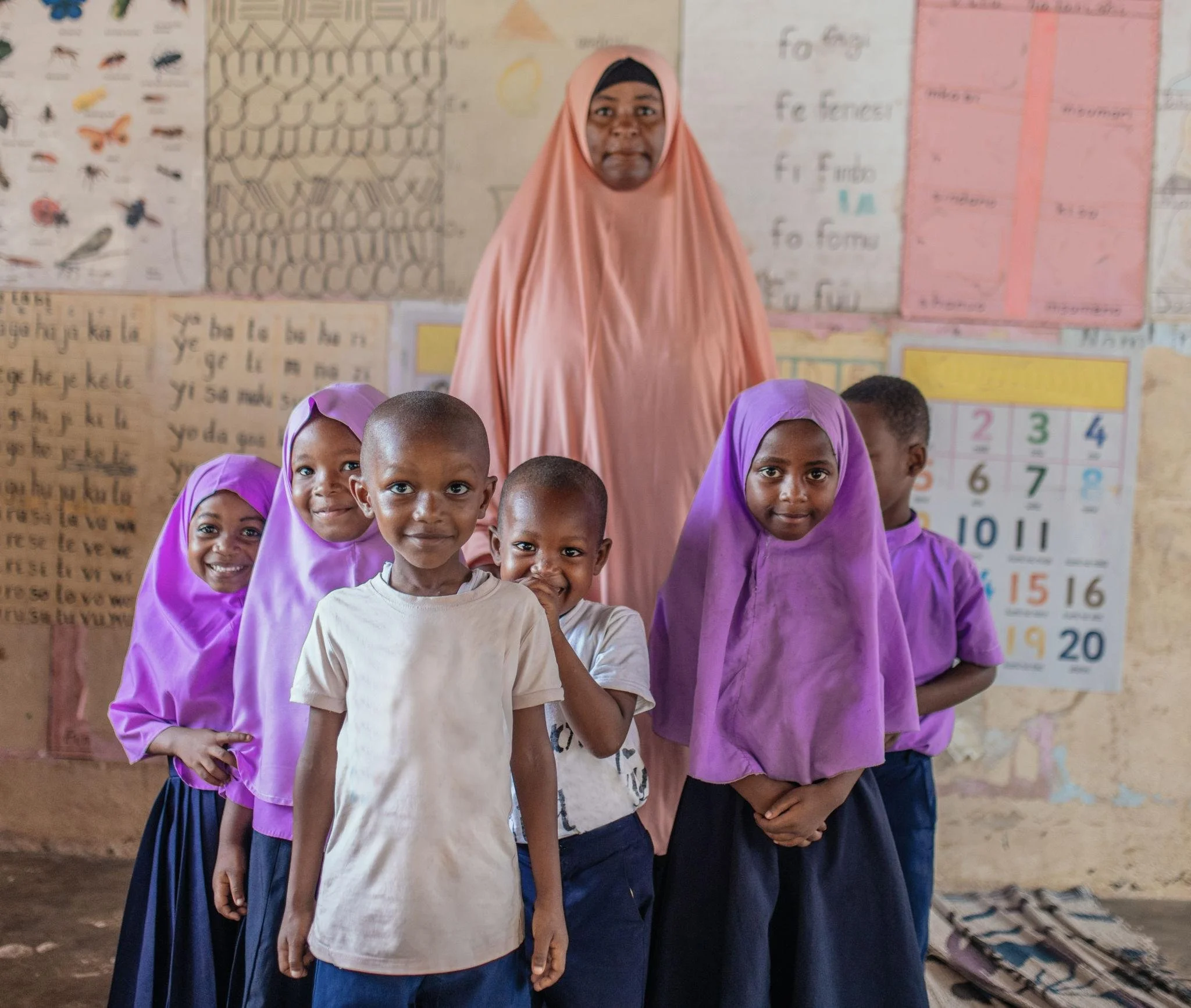 A woman wearing a peach-colored hijab and several children with purple headscarves standing in a classroom with educational posters on the wall.