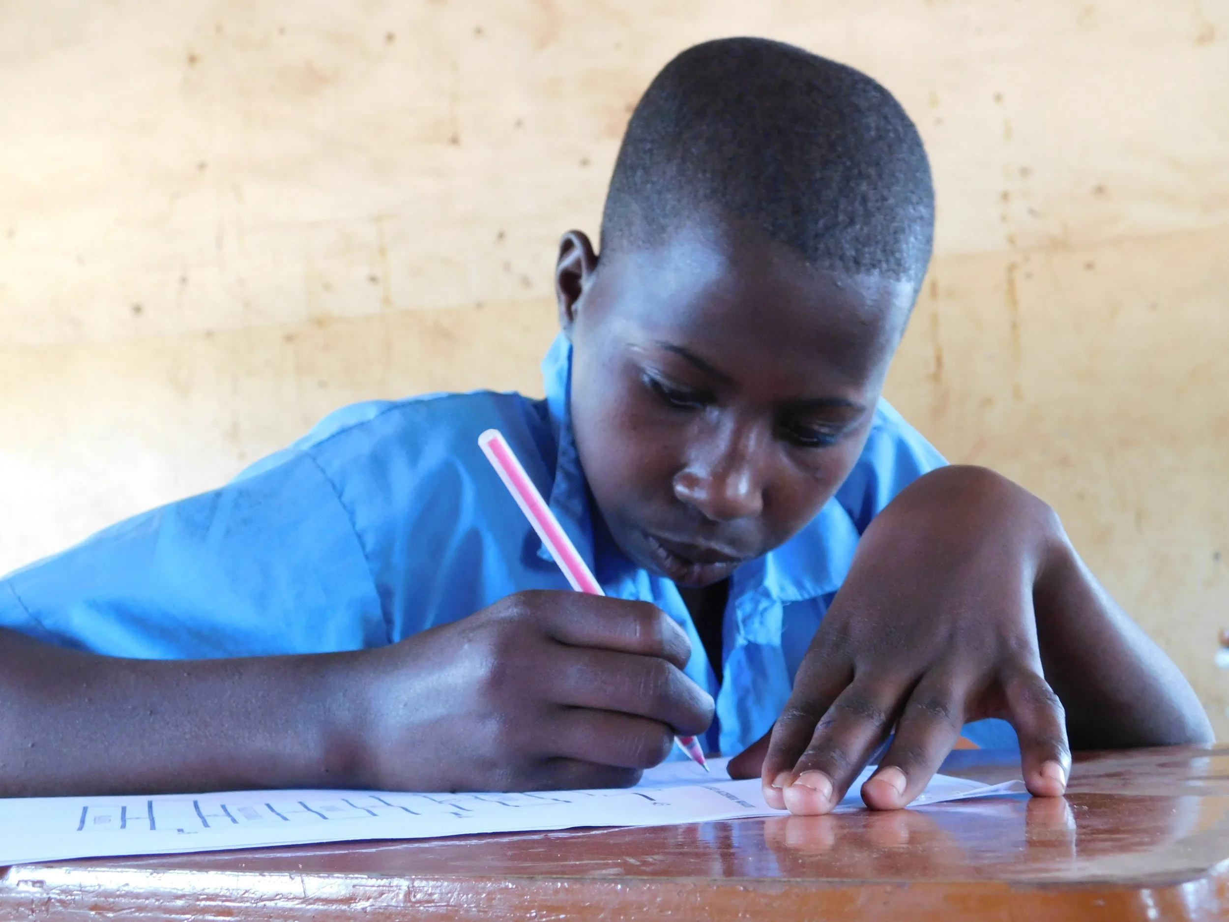 A student in a blue uniform writing at a classroom desk.