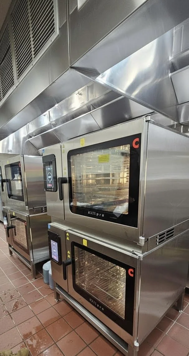Commercial kitchen with multiple stainless steel ovens and appliances, tiled floor, and ventilation system overhead.