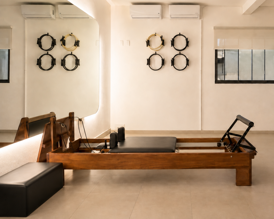 Sala de pilates con un marco en forma de cama de madera, una almohadilla en el centro, pesas de manos, tapete negro, yoga mat, muchos auriculares en la pared y aire acondicionado.