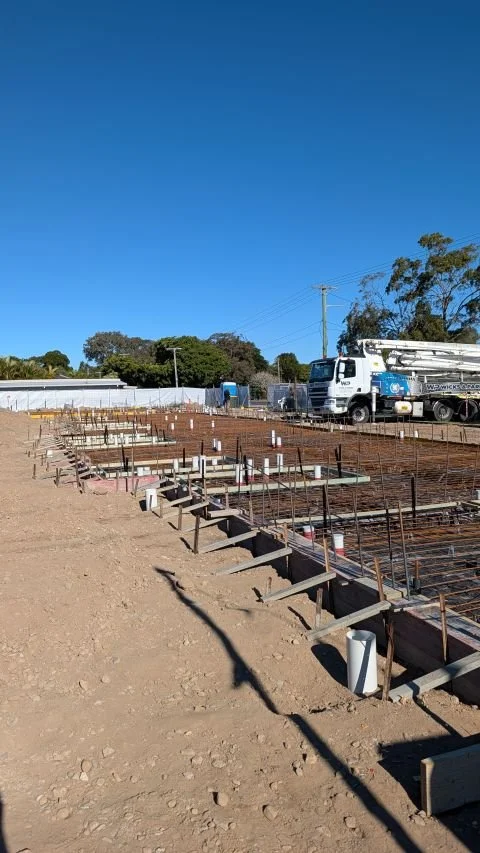 Slab inspection at Club Iluka new Motel mid construction. A live construction site with steel rebar framework for a concrete foundation, a large truck parked nearby ready to pour concrete, and blue skies overhead.