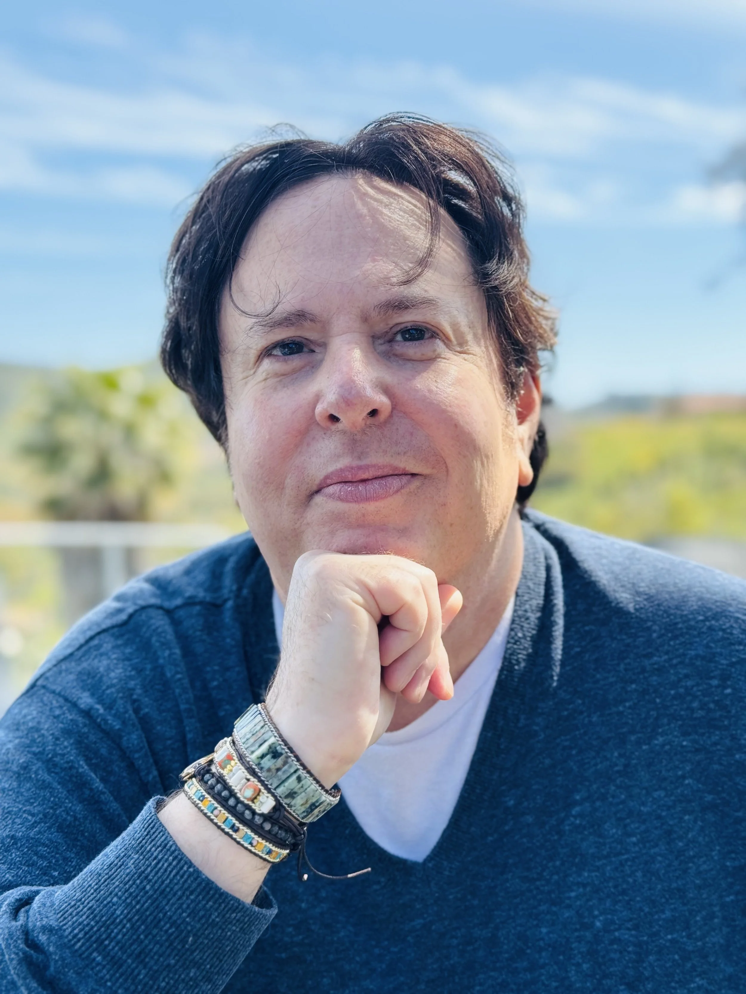 A male real estate agent with dark hair and light skin posing outdoors against a southern California background of sky and greenery, wearing a blue sweater, white undershirt, and decorative bracelets on his wrist.