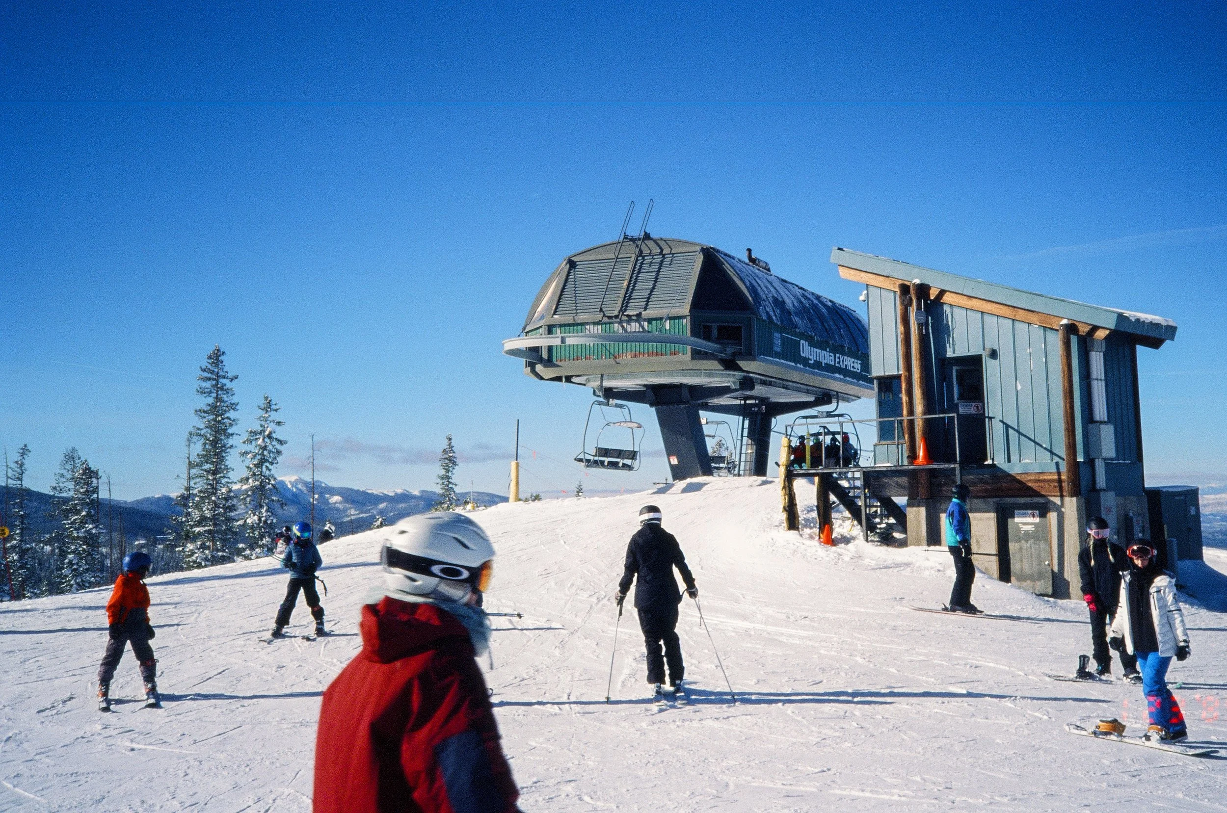 A snowy ski resort with skiers and snowboarders, a chairlift station, and mountainous landscape in the background under a clear blue sky.