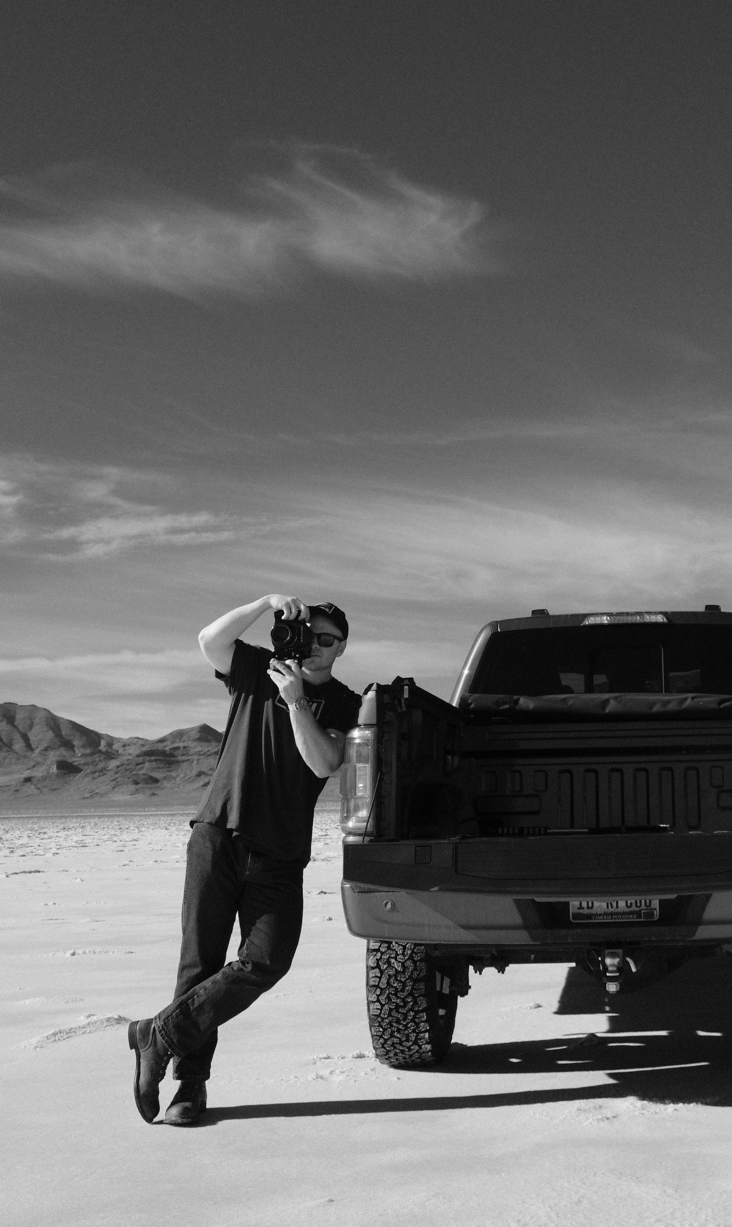 A man wearing sunglasses, a baseball cap, and casual clothing is leaning against the back of a pickup truck in a desert landscape, holding a camera and taking a photo.