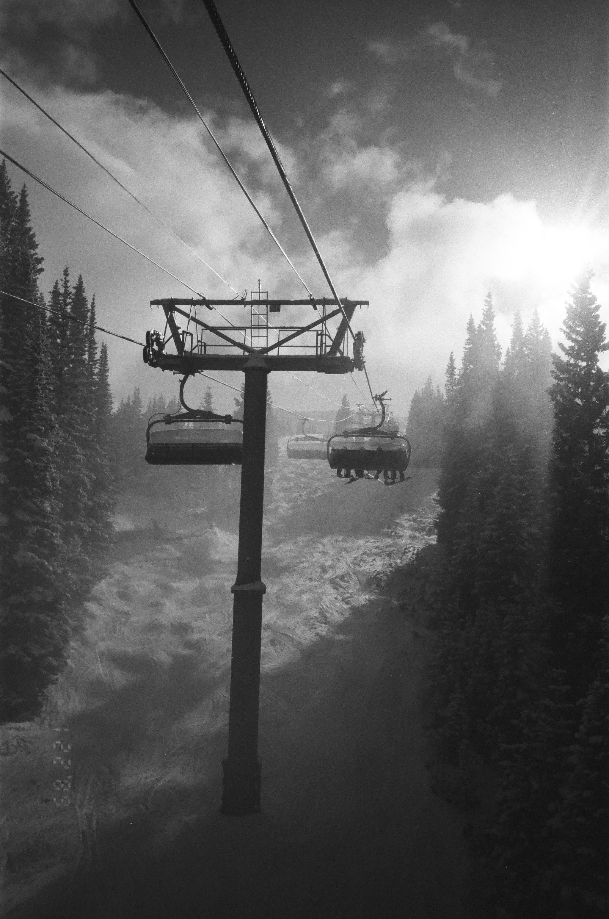 Black and white photo of a ski lift in a snowy mountainous forest with trees and cloudy sky.