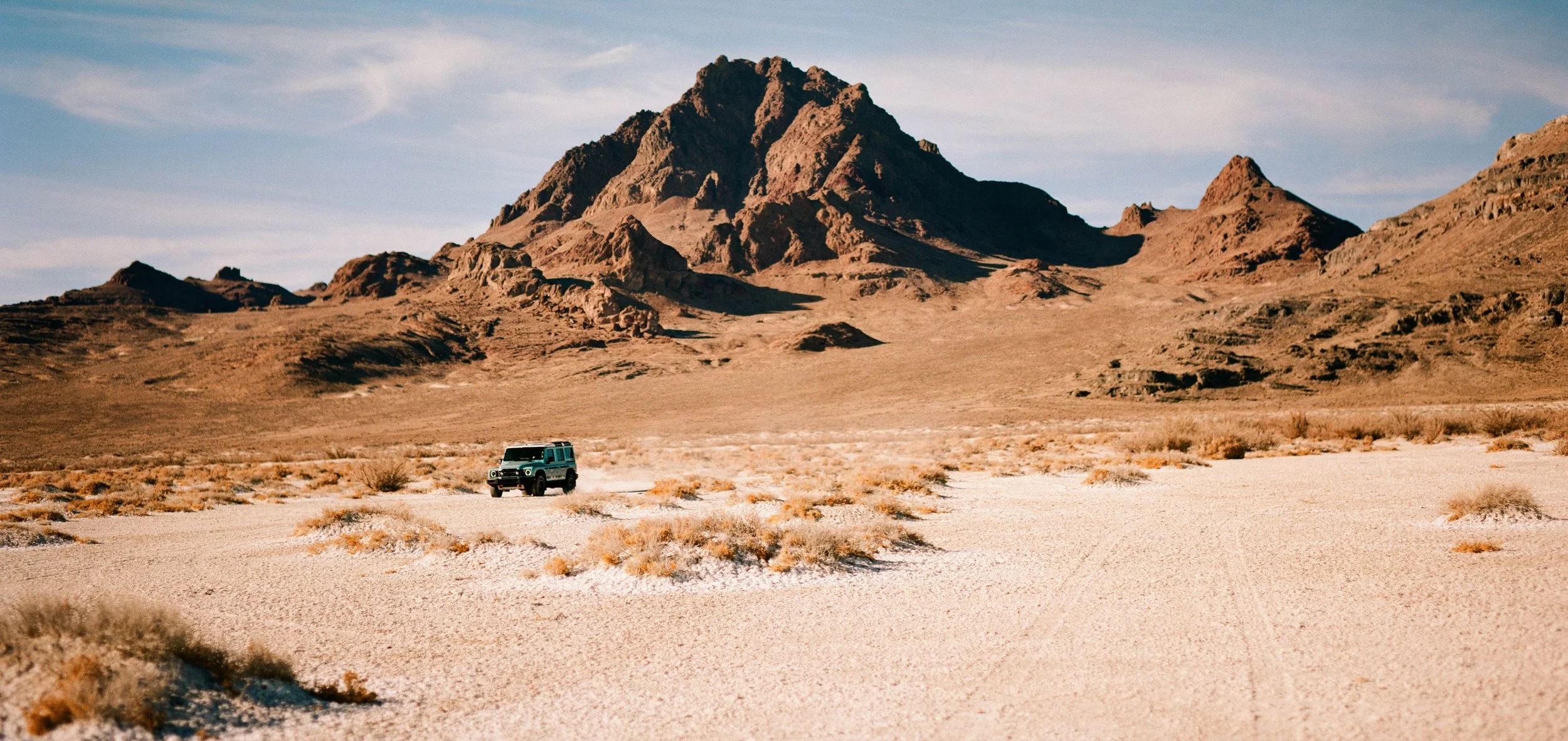 A vehicle driving through a desert landscape with mountains in the background under a partly cloudy sky.