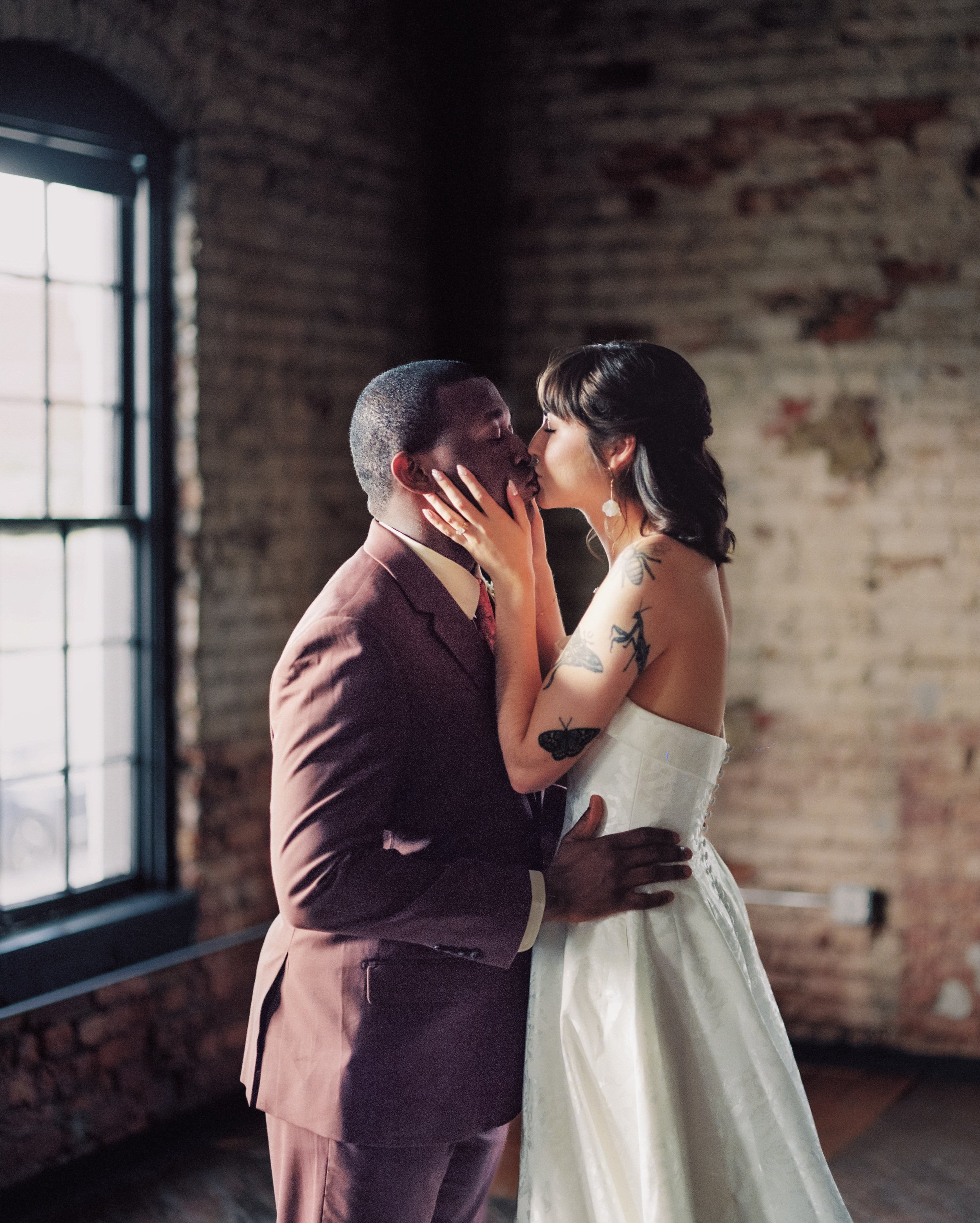 A couple shares a kiss inside a room with exposed brick walls and large window, with natural light illuminating them.