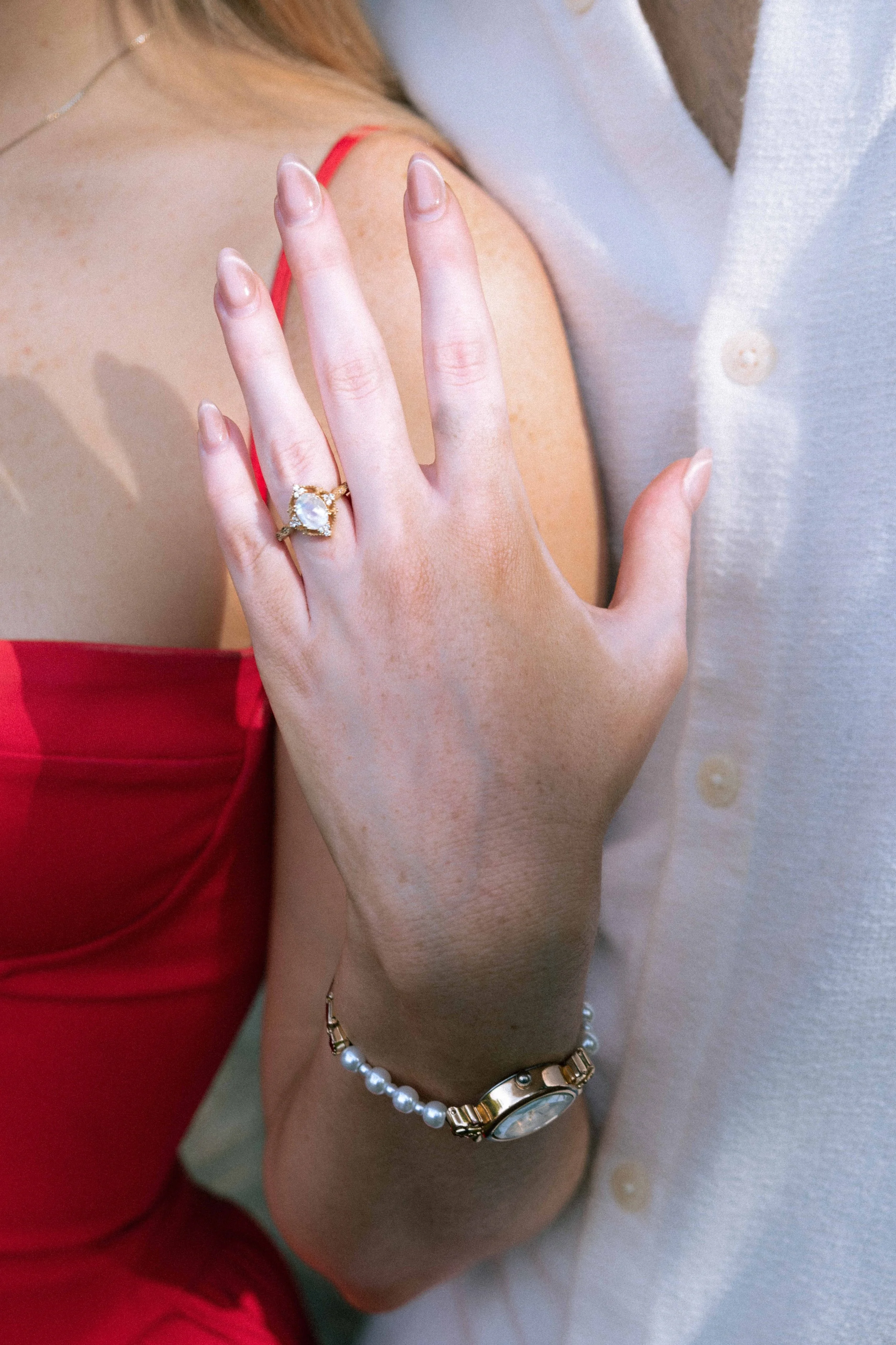 Close-up of a woman wearing a red dress and jewelry, touching another person's arm, with focus on her hand and a ring on her finger.