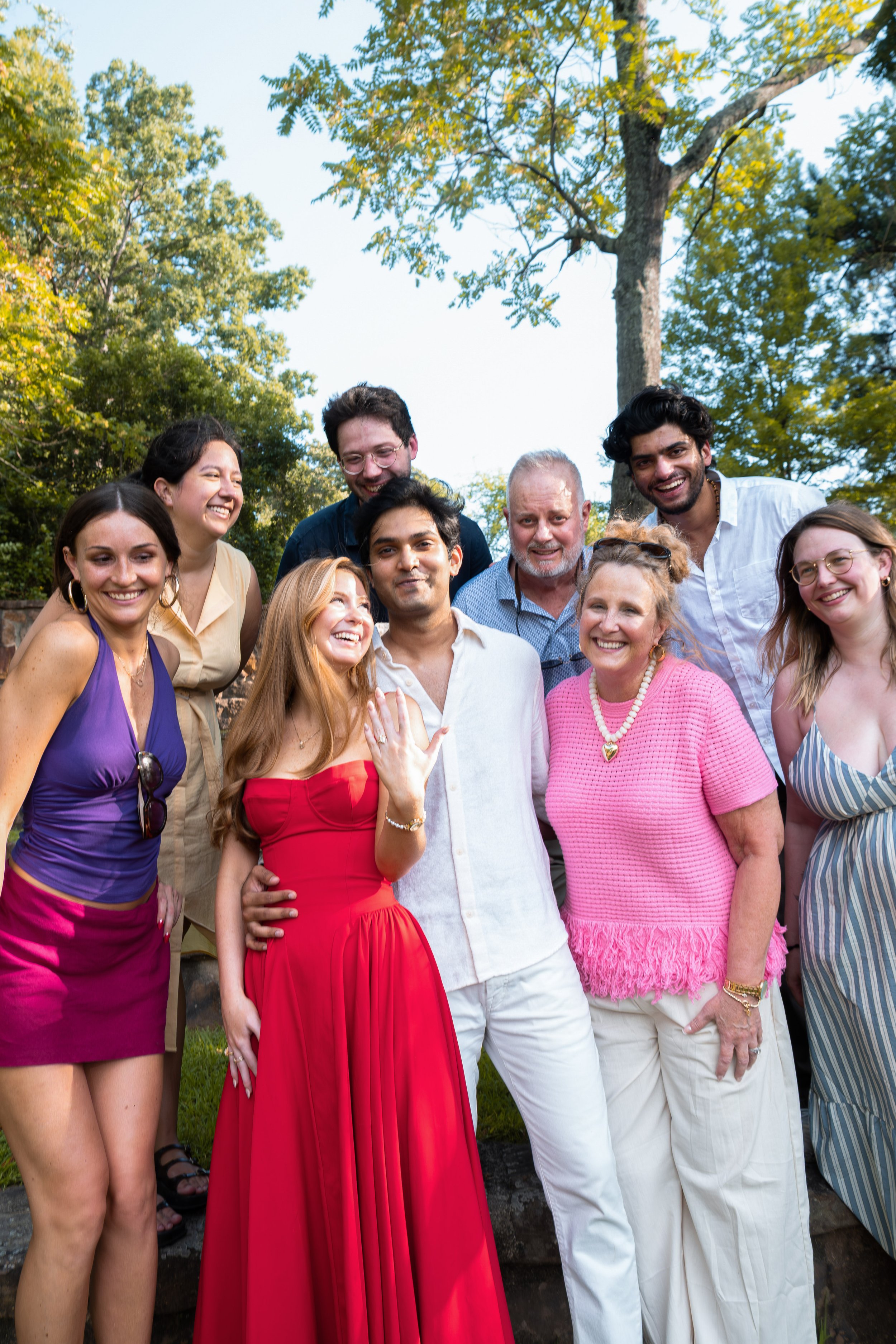 A diverse group of people smiling and posing outdoors in a park with trees in the background.