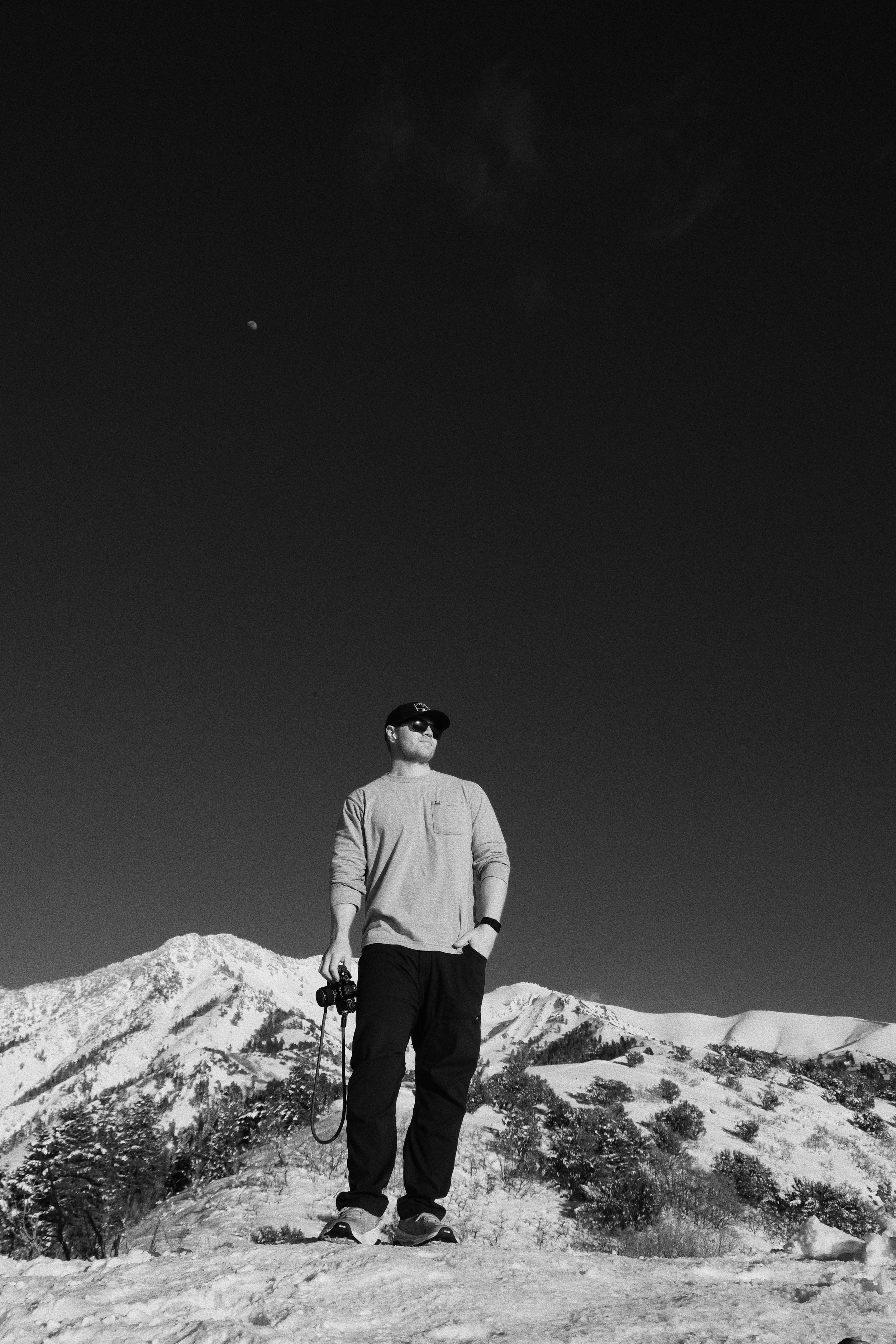 A man standing on snow-covered ground with snowy mountains in the background, holding a camera strap, wearing sunglasses, a cap, and casual winter clothing, under a dark sky with a visible moon.
