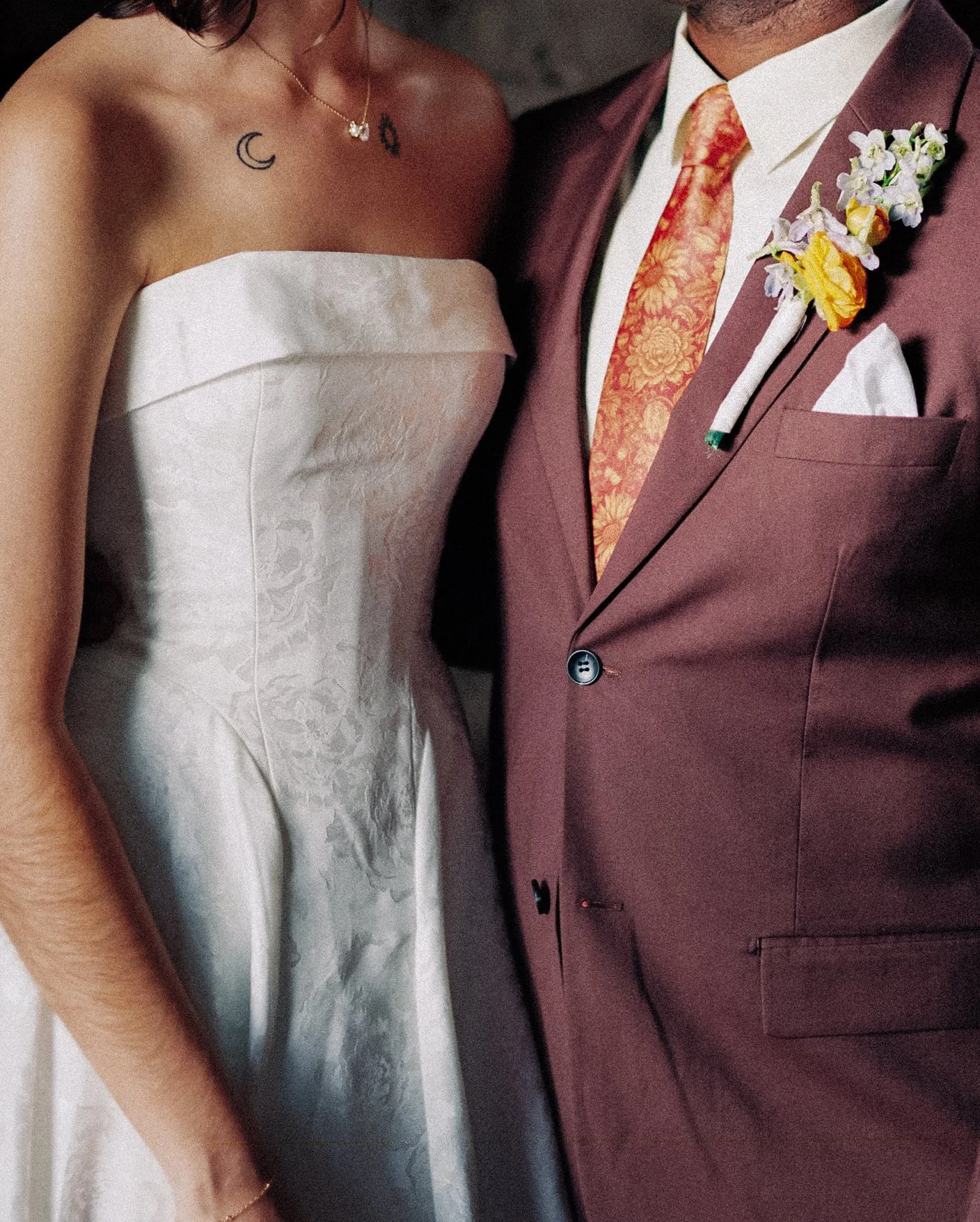 A close-up photo of a bride and groom at a wedding. The bride is wearing a strapless white wedding gown, a delicate necklace, and has small tattoos on her chest. The groom is dressed in a maroon suit with a floral-patterned tie, a white shirt, and a matching boutonnière with flowers pinned to his lapel.