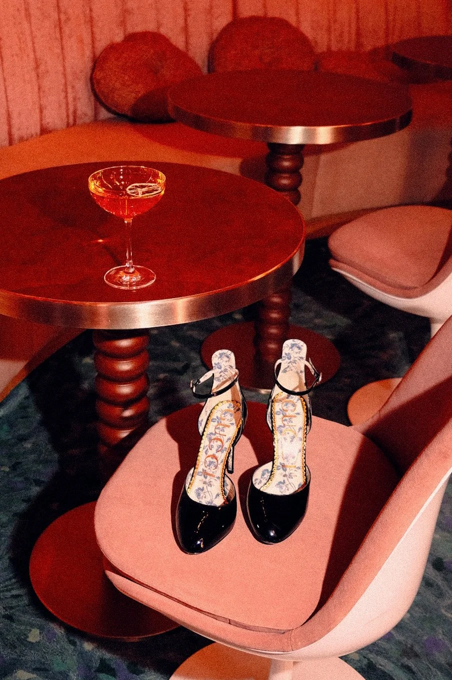 Black high-heeled shoes with ankle straps placed on a pink upholstered chair in a dimly lit room with wooden tables and a dark patterned carpet.