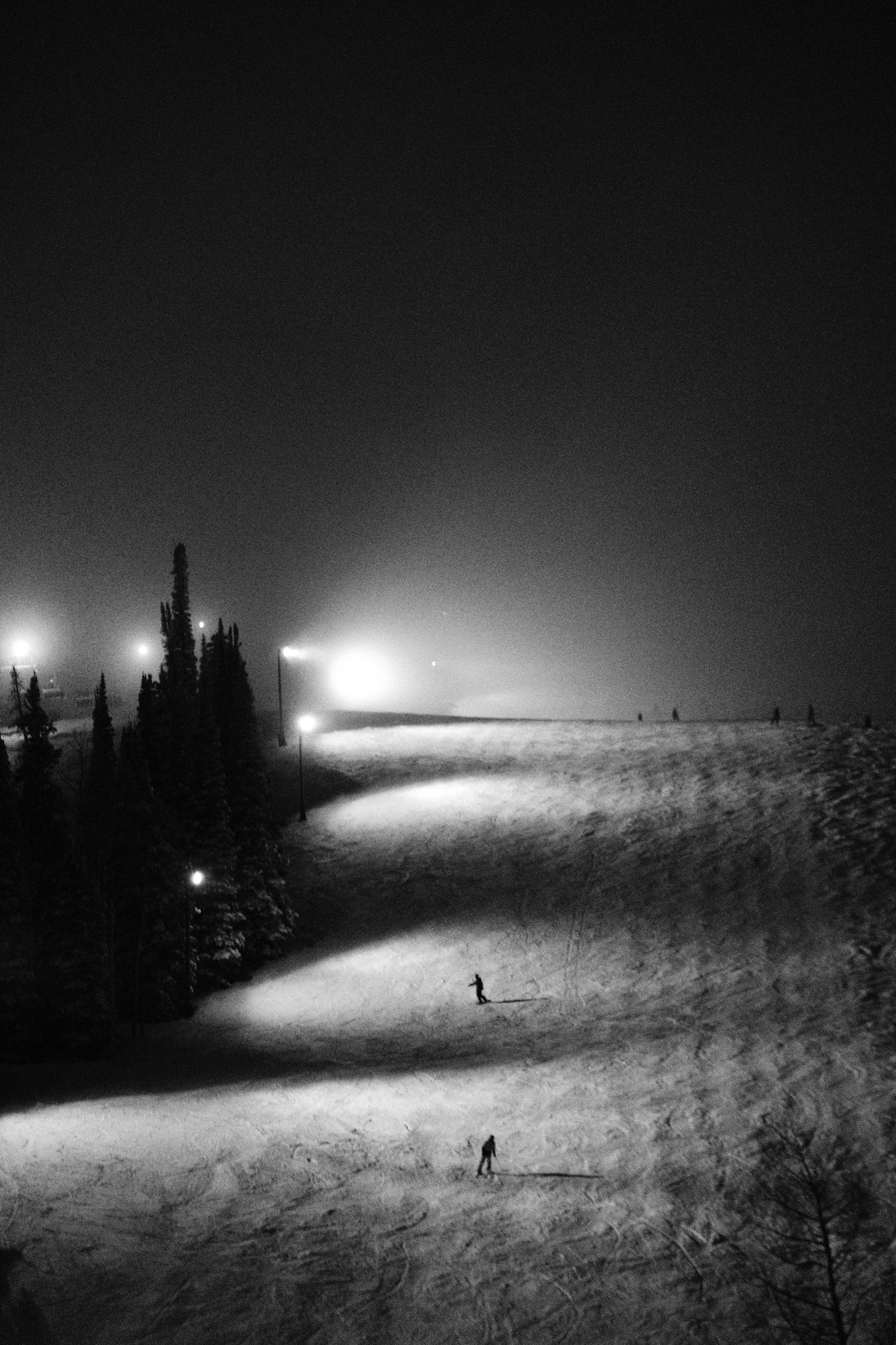 Nighttime view of a snowy landscape with a few people skiing, illuminated by several bright lights, with trees silhouetted against the dark sky.