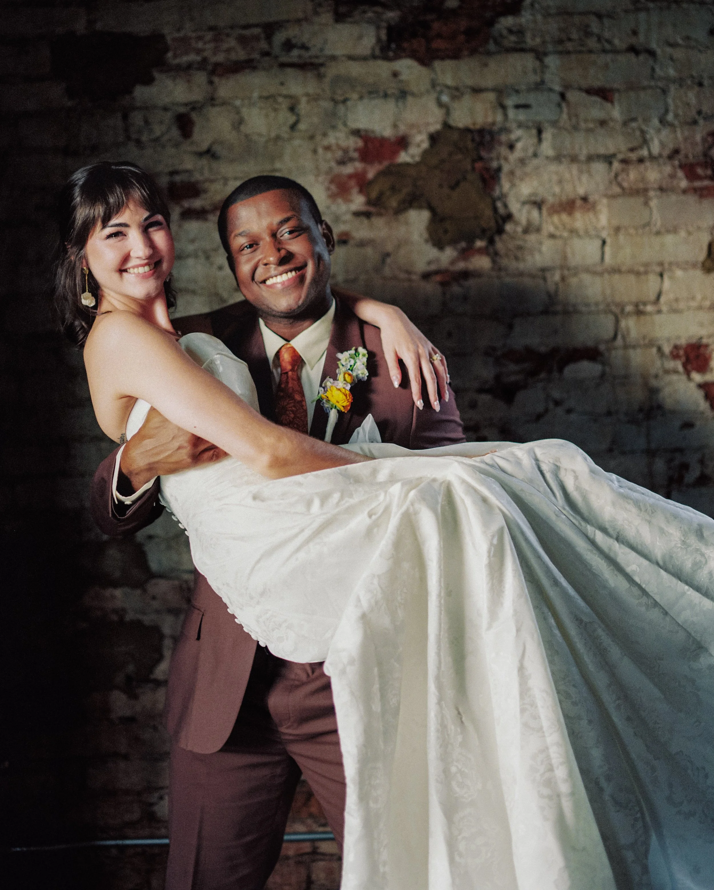 A smiling couple, a woman with dark hair and a man with short hair, celebrating with a hug. They are dressed in formal attire, with the man wearing a suit and tie with floral decorations. The background features a rustic brick wall.