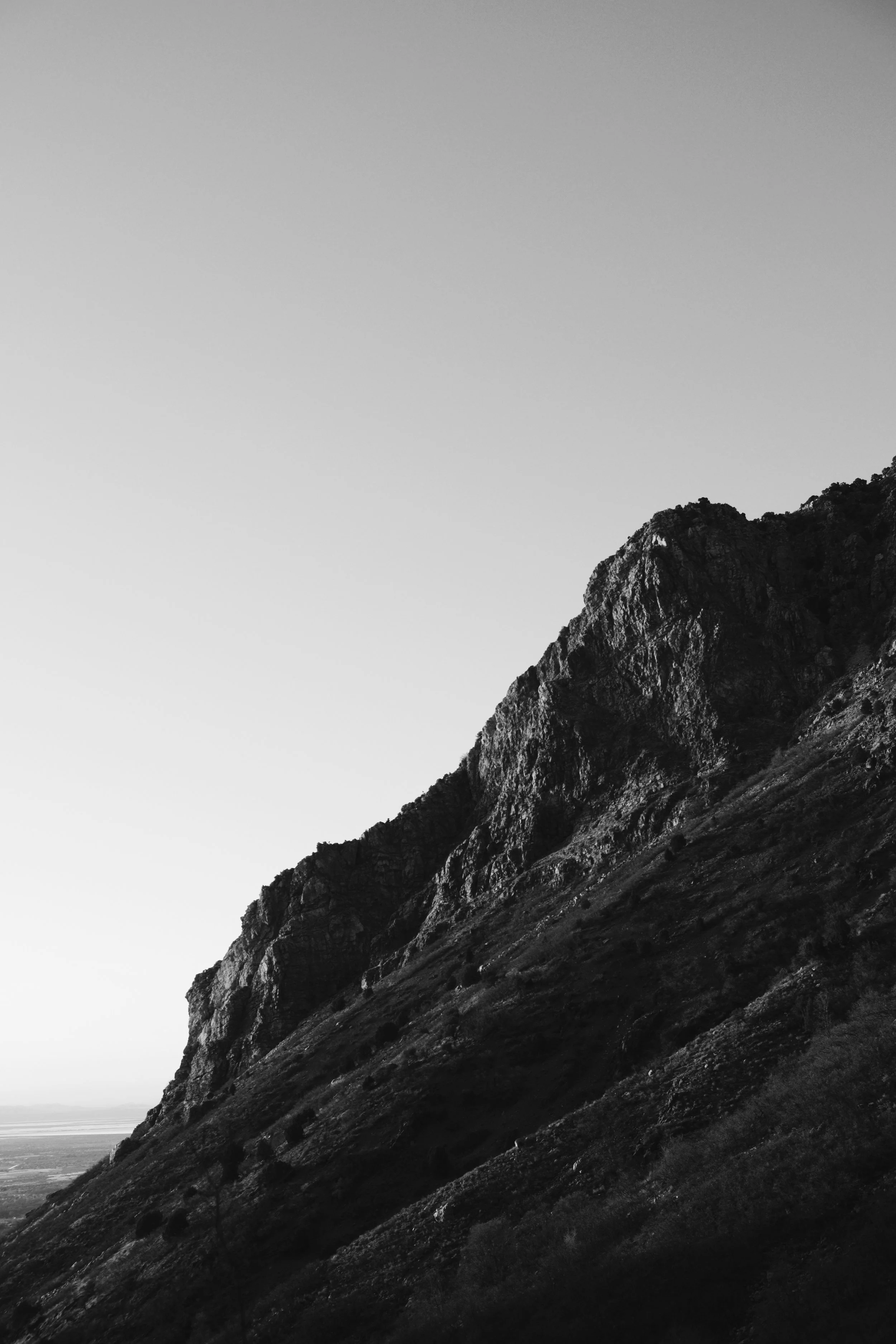 Black and white photo of a steep mountain slope with rocky terrain and sparse vegetation, with a clear sky above.
