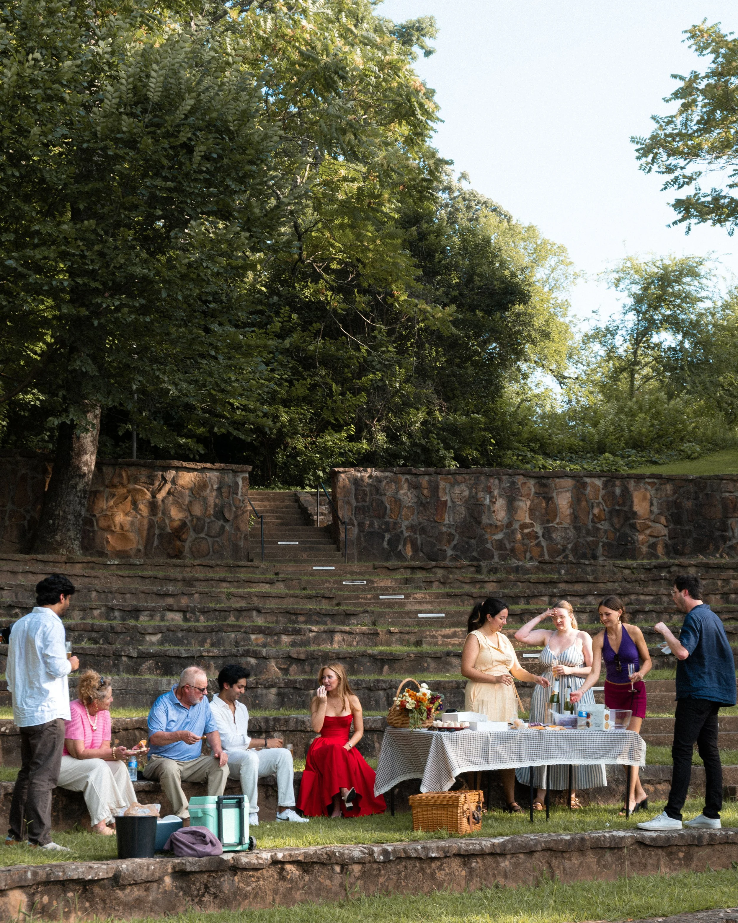 A group of people gathered at an outdoor event on stone steps, with trees and a stone wall in the background. Some are sitting, and some are standing near a table with food and drinks.