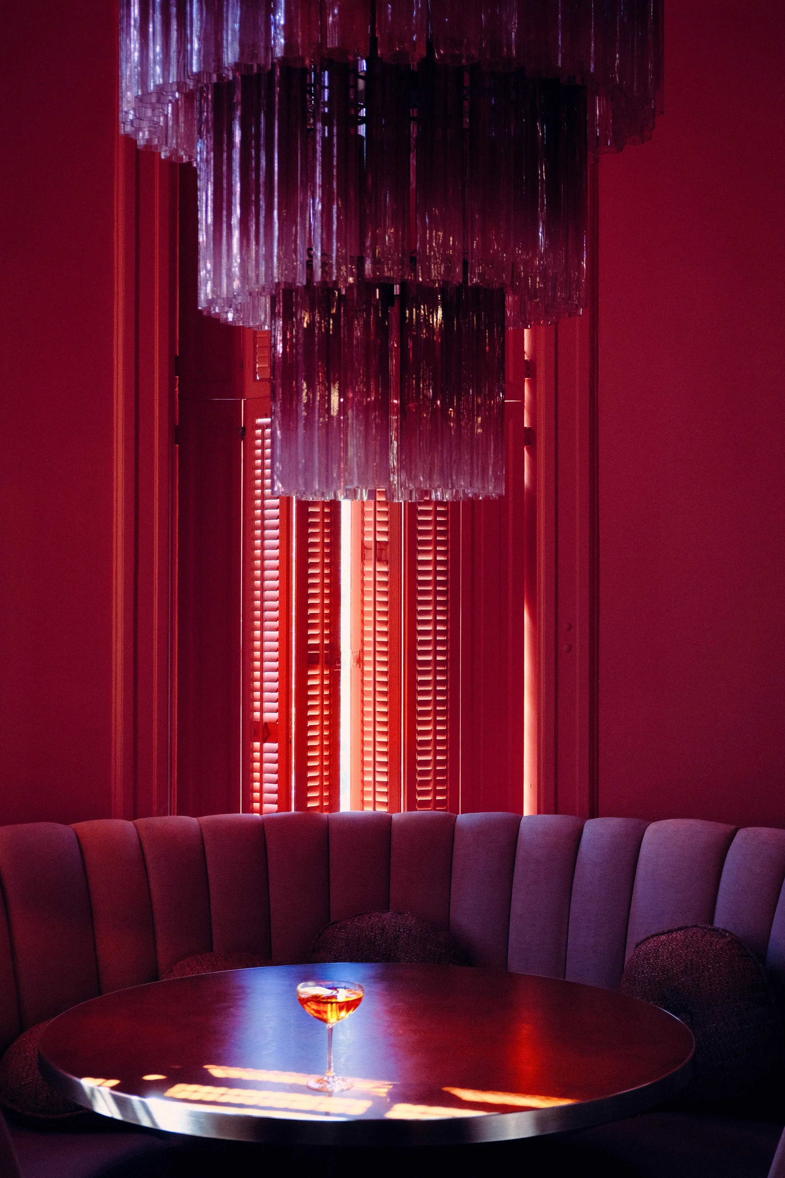 A dimly lit room with a round table in the foreground holding a glass of amber-colored drink. Behind the table is a curved, upholstered purple seating area, and on the wall, there are closed pinkish shutters. Above the shutters, a large modern chandelier with hanging rectangular glass or acrylic pieces reflects some light.