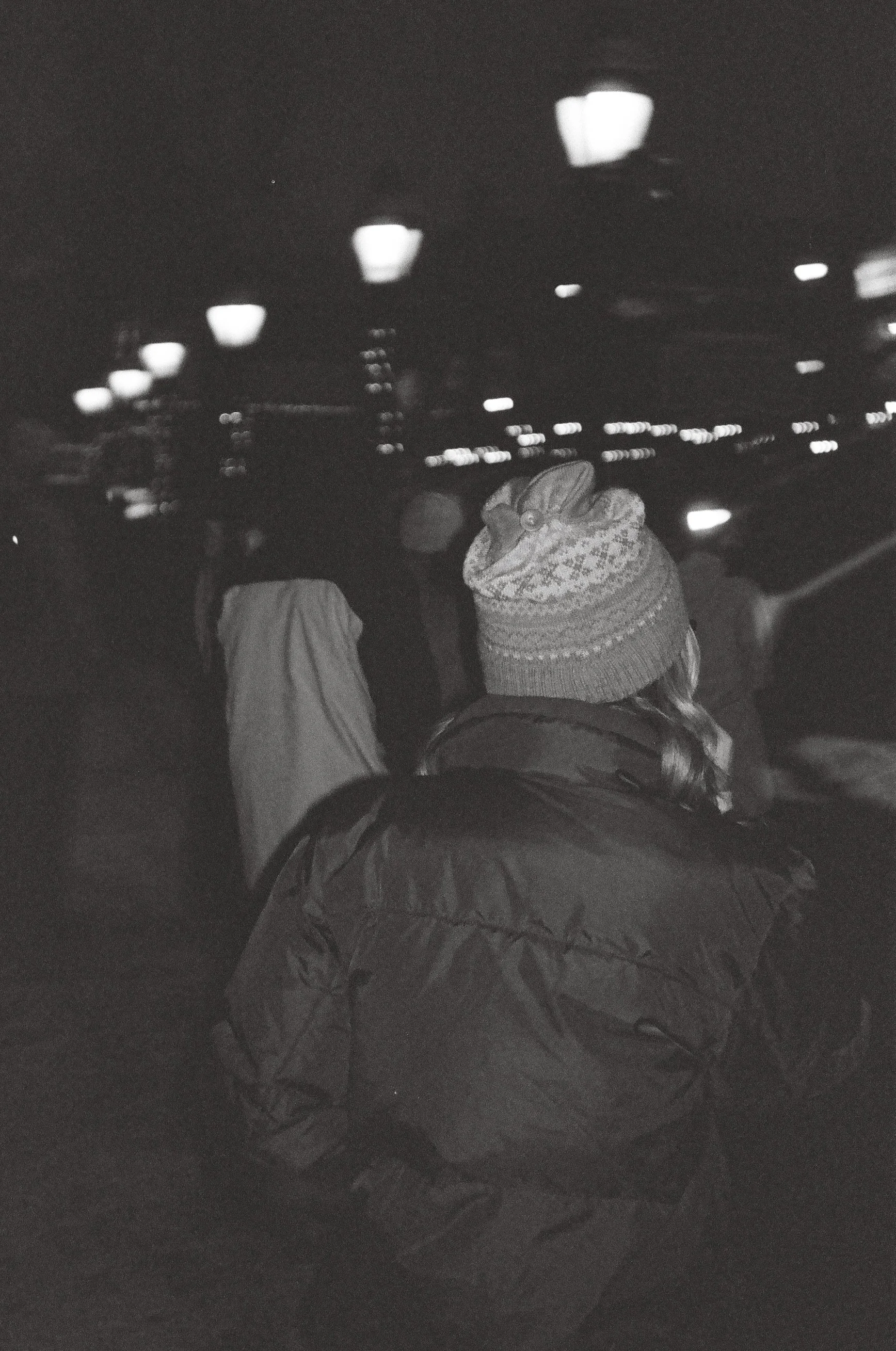 Nighttime scene inside a building with illuminated ceiling lights, showing a person with a colorful knit hat and a coat, standing in front of a reflective surface or window.