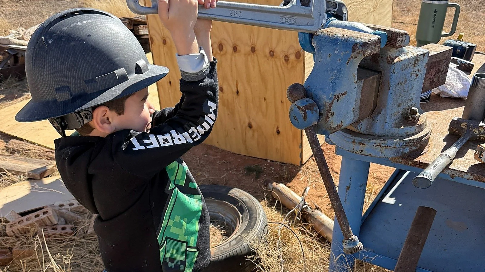A young boy wearing a safety helmet is working with a large metal vise attached to a workbench outdoors, possibly at a construction or woodworking site.