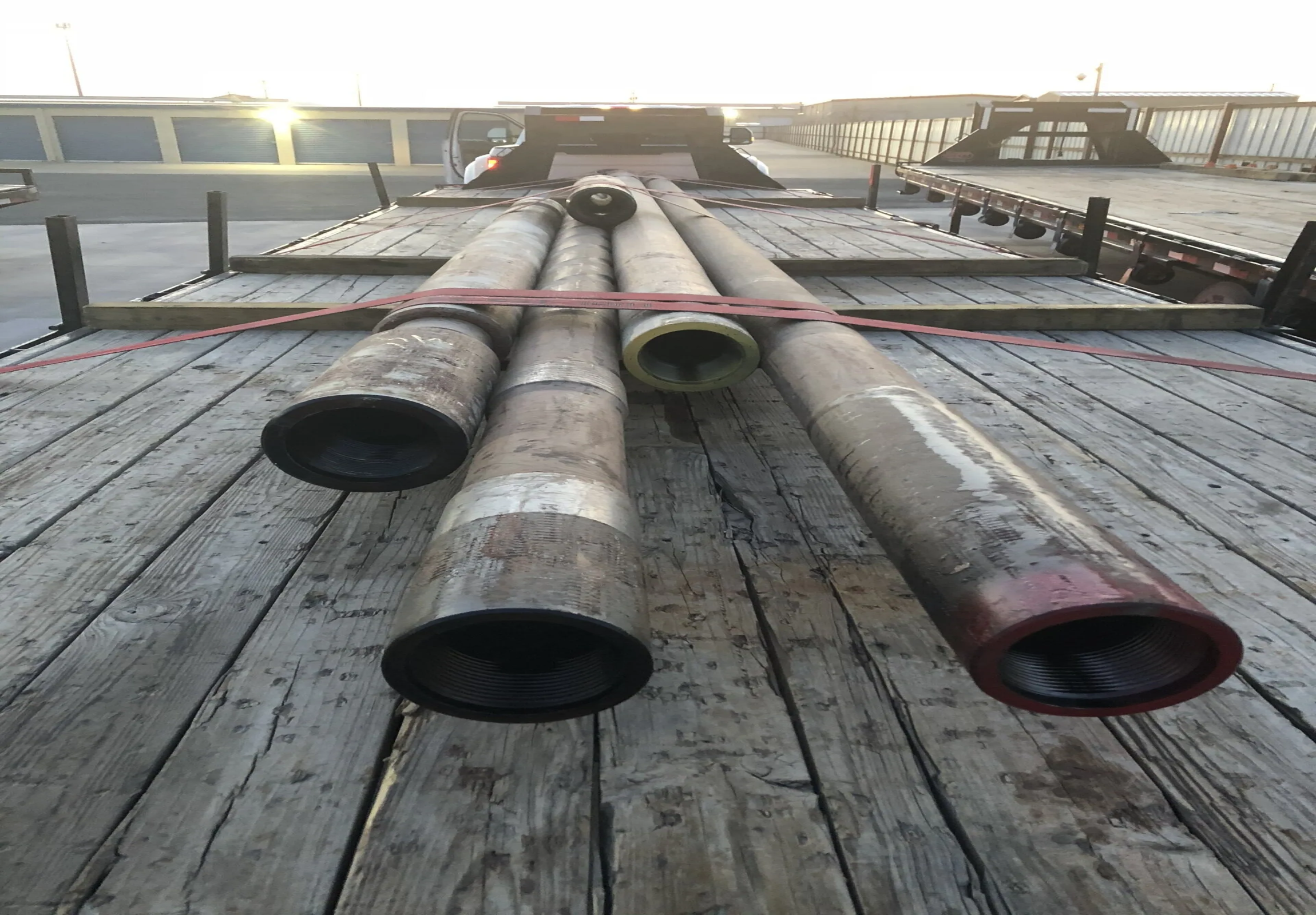 A flatbed truck transporting large industrial pipes on a wooden platform with a white car in the background.