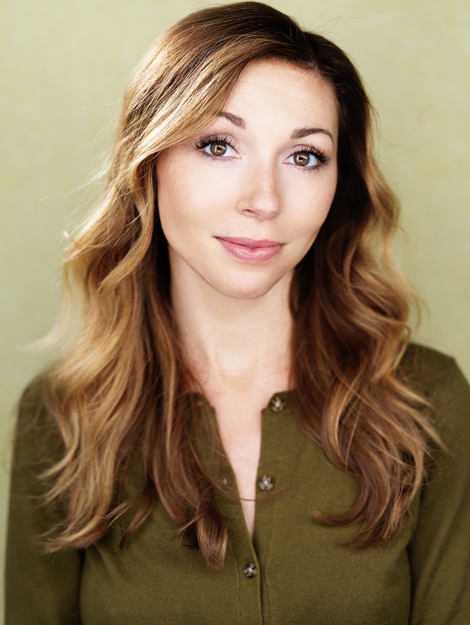 Close-up portrait of a young woman with light brown hair, hazel eyes, and a subtle smile, wearing an olive green button-up top, against a soft green background.