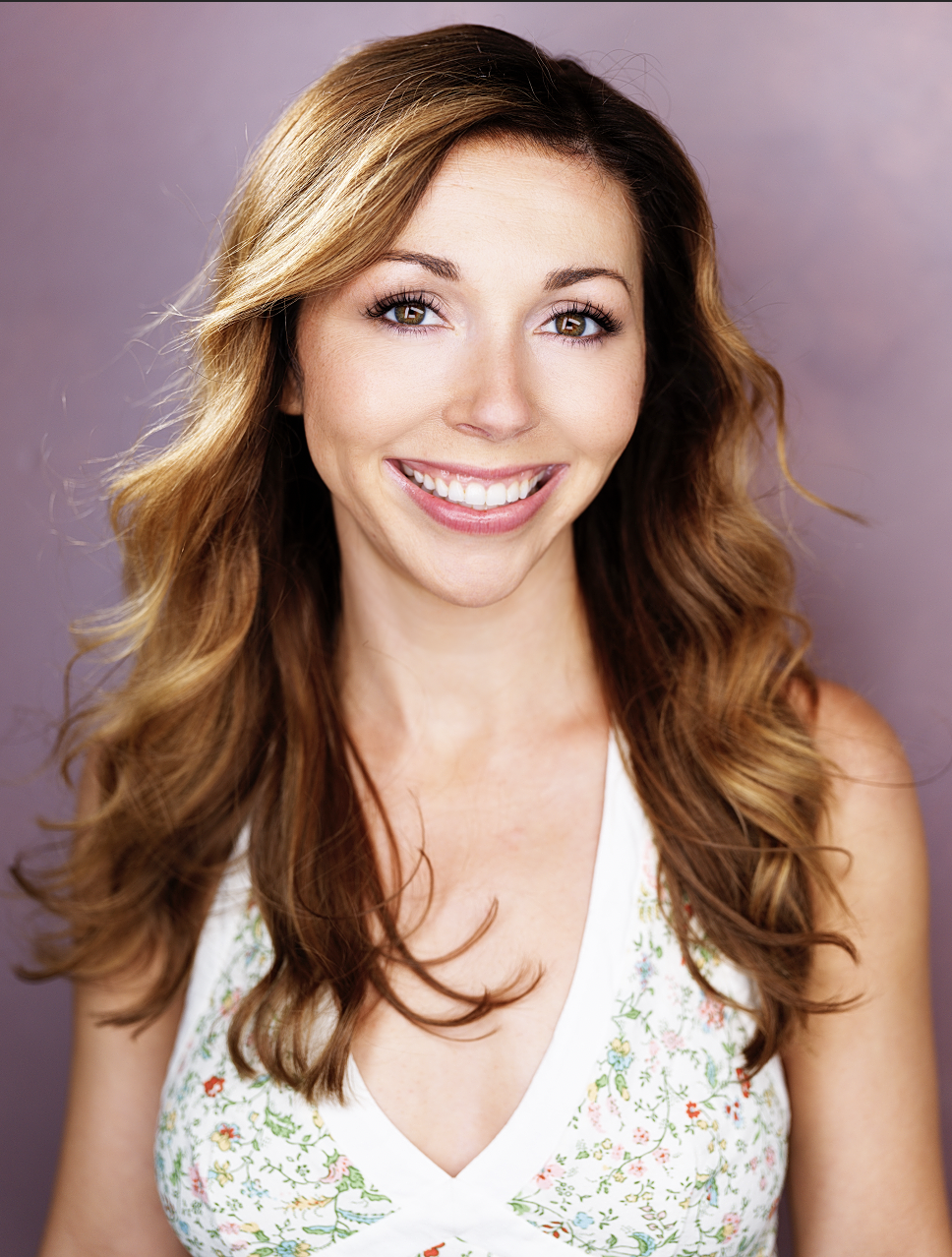 Portrait of a young woman with wavy, reddish-brown hair, smiling, wearing a white floral dress, against a purple background.