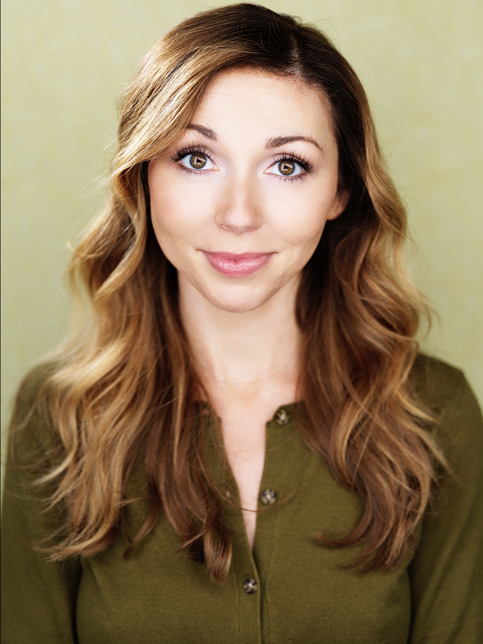 A woman with long, wavy, light brown hair and hazel eyes smiling at the camera, wearing an olive-green top with buttons, against a light green background.