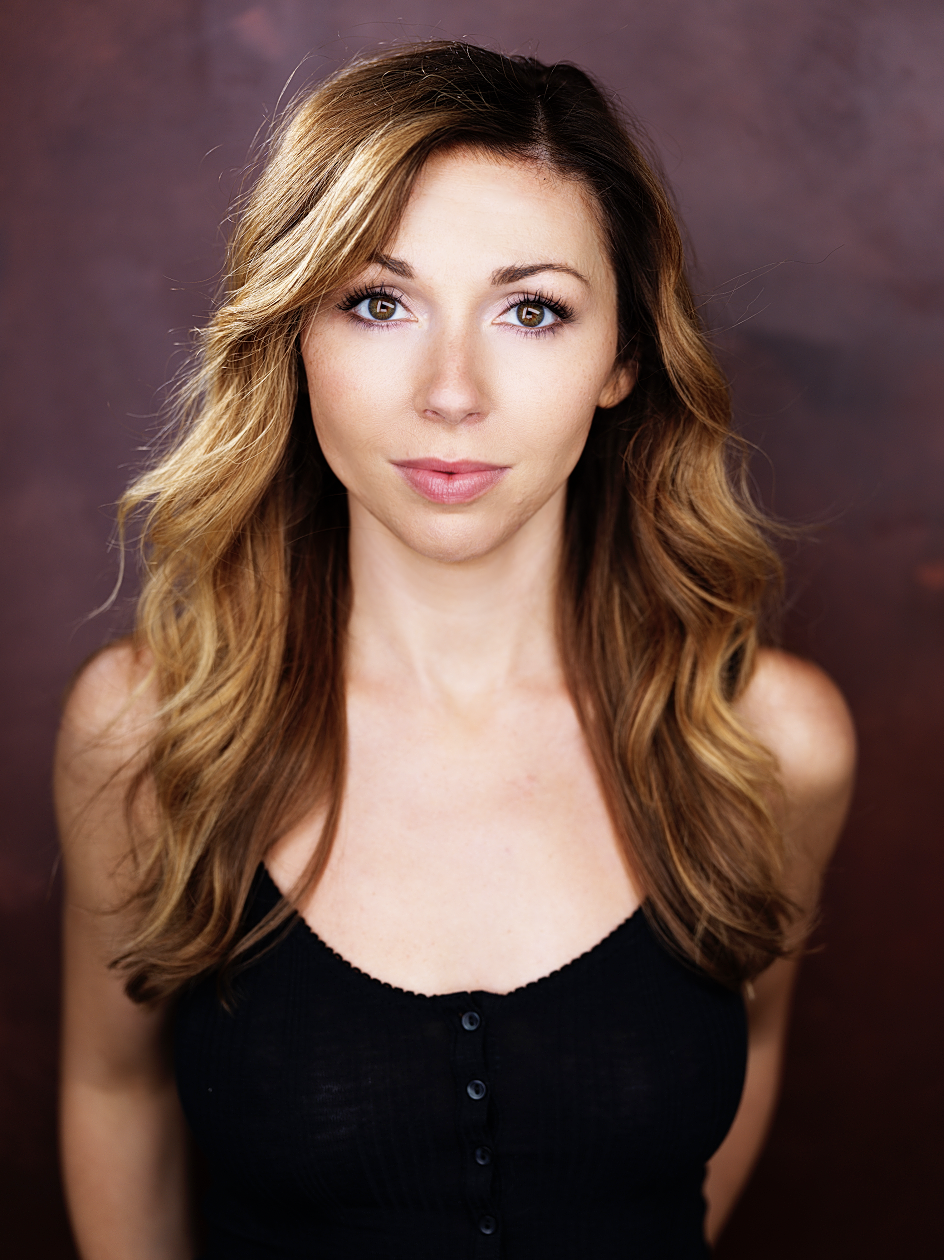 A woman with long, wavy, light brown hair and hazel eyes wearing a black sleeveless top, looking directly at the camera against a blurred, dark background.