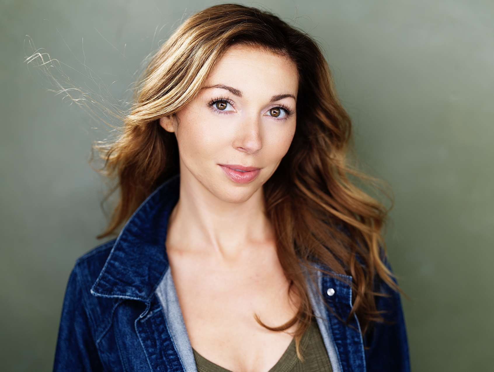 A woman with long, wavy brown hair and fair skin, wearing a denim jacket over a green shirt, standing against a muted green background.